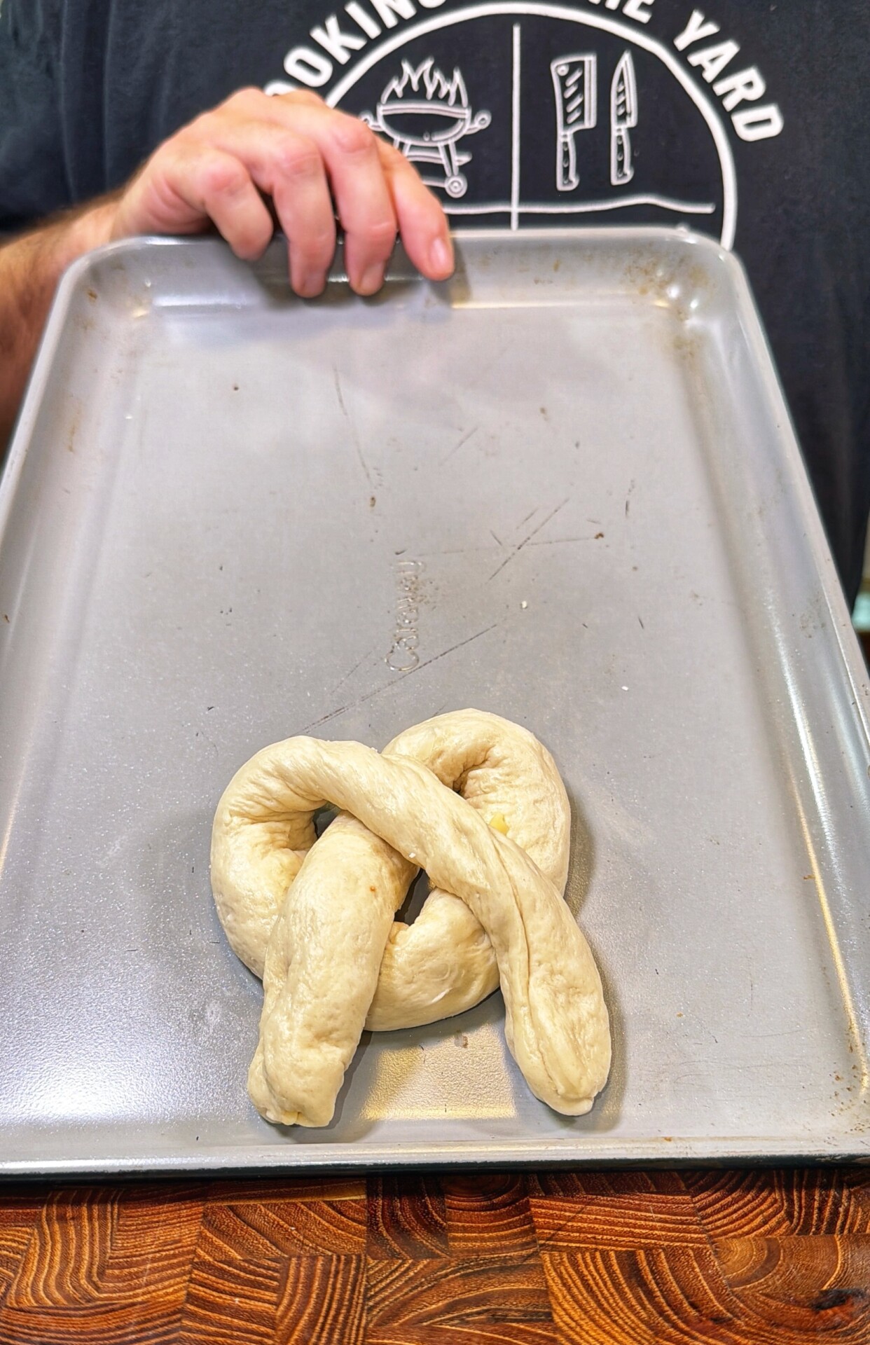 A hand holds a baking tray with an uncooked pretzel-shaped dough on it. The person is wearing a black shirt with a white cooking-themed graphic on the chest.