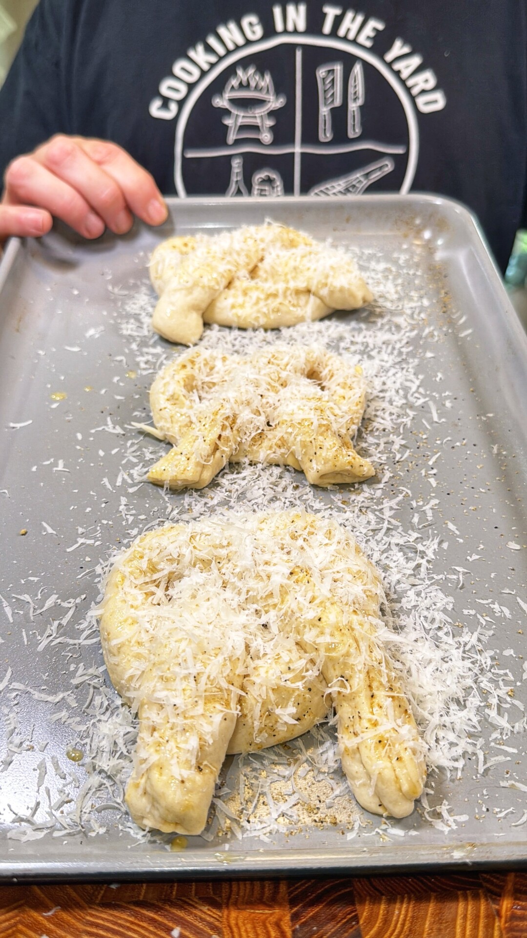 A person holds a baking tray with three unbaked, bunny-shaped bread dough pieces topped with grated cheese. The person is wearing a black Cooking in the Yard shirt.