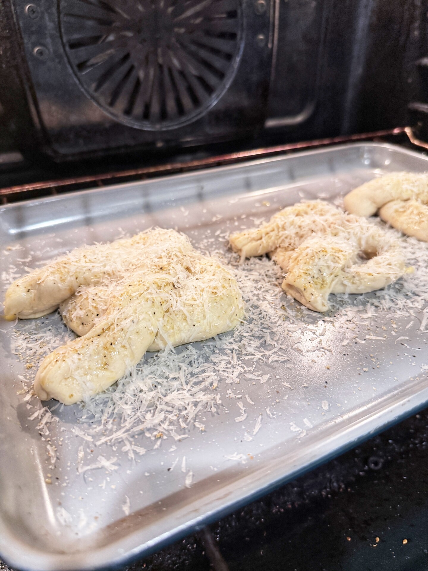 Two pieces of uncooked dough shaped like hands, topped with shredded cheese and herbs, sit on a metal baking sheet inside an oven.