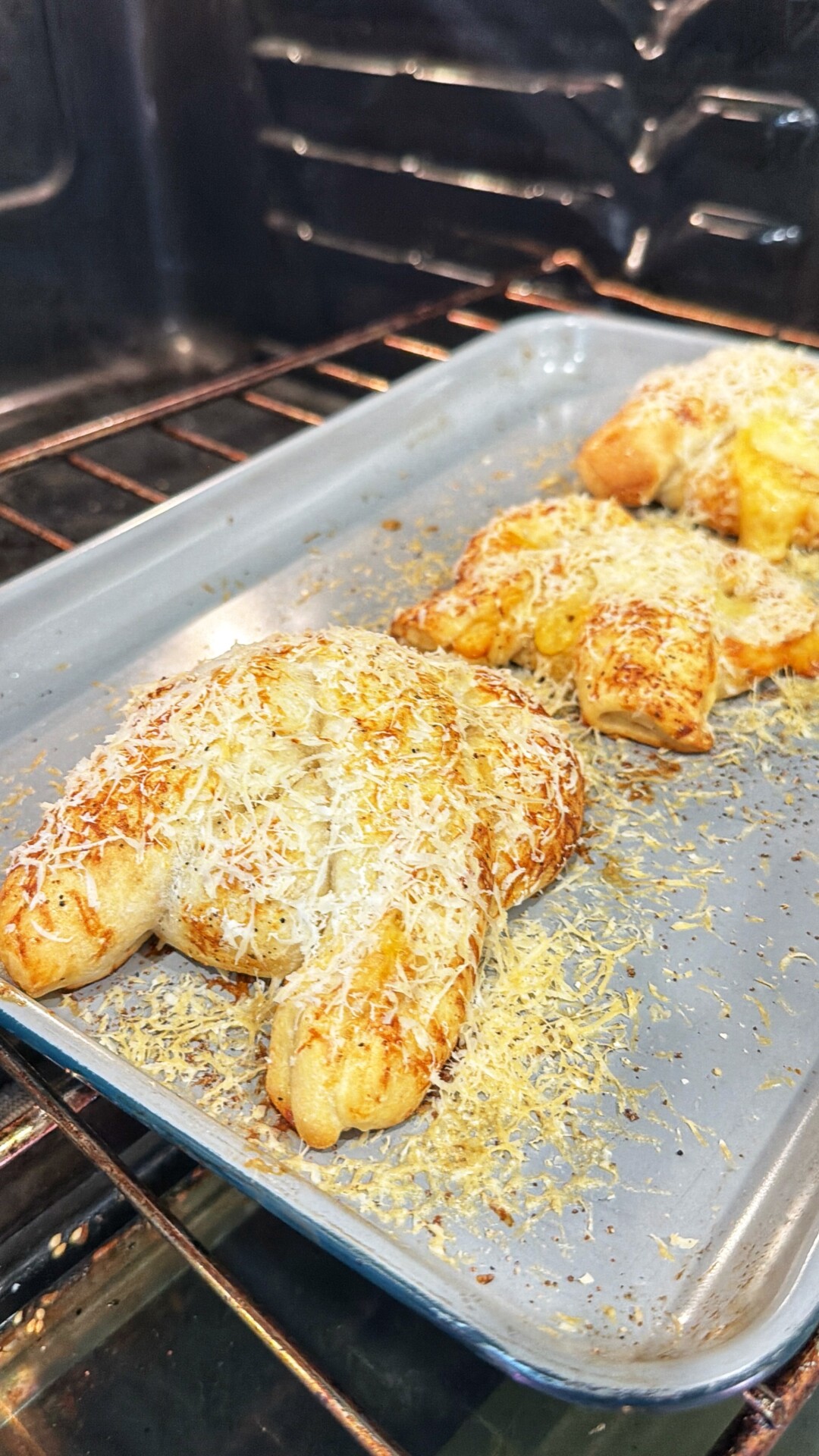 Golden, cheese-covered pastries baking on a metal tray inside an oven. The pastries are crescent-shaped and sprinkled with shredded cheese, with the oven racks visible in the background.