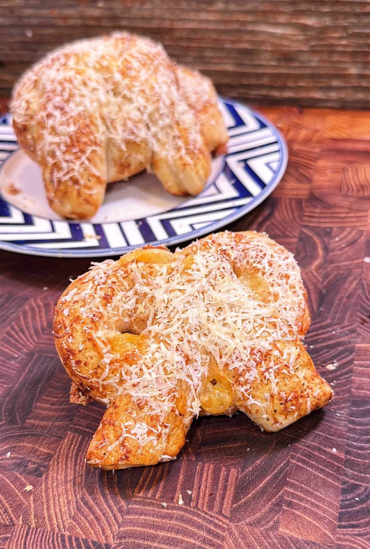 Two soft pretzels topped with grated cheese, one on a patterned plate in the background and one on a wooden surface in the foreground.
