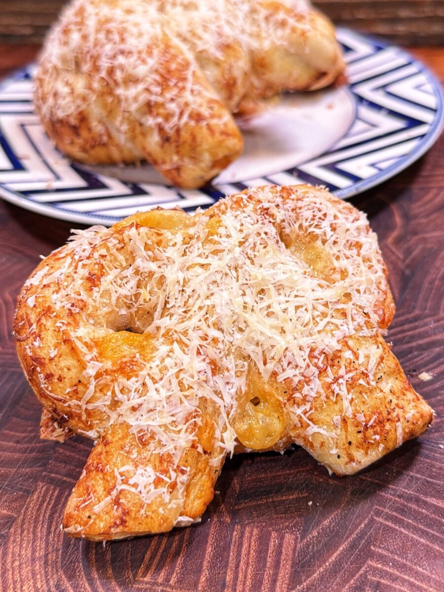 A golden, flaky pastry topped with grated cheese sits on a wooden board, with another pastry on a patterned plate in the background.