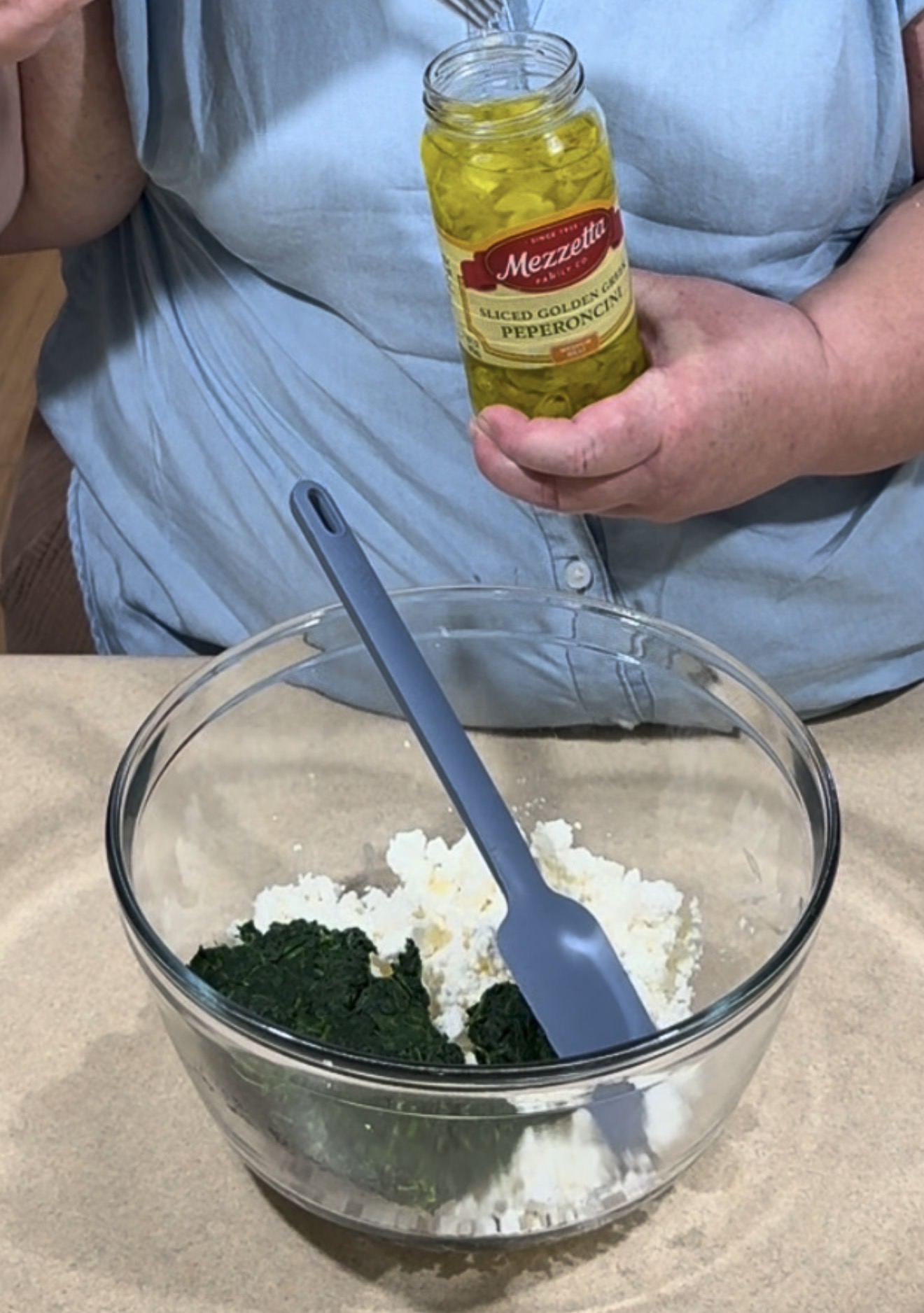 A person holds a jar of sliced golden Greek pepperoncini over a glass bowl containing spinach and cheese with a gray spatula inside.