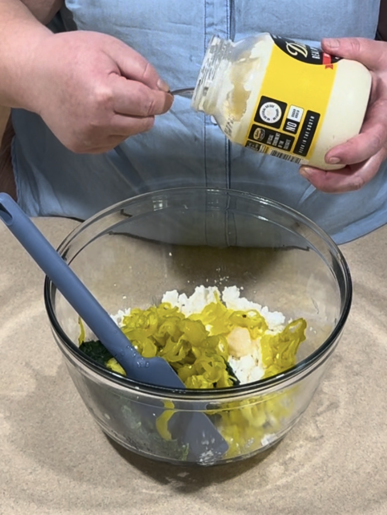 A person scoops mayonnaise from a jar into a glass bowl filled with sliced pickles and other ingredients. A blue spatula rests in the bowl.