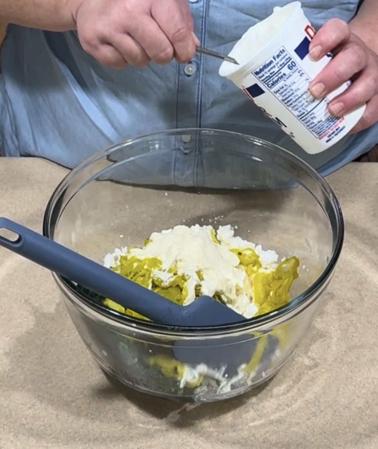 A person in a blue shirt scoops sour cream from a container into a glass bowl with mayonnaise and seasonings, using a spoon. A blue spatula rests inside the bowl.