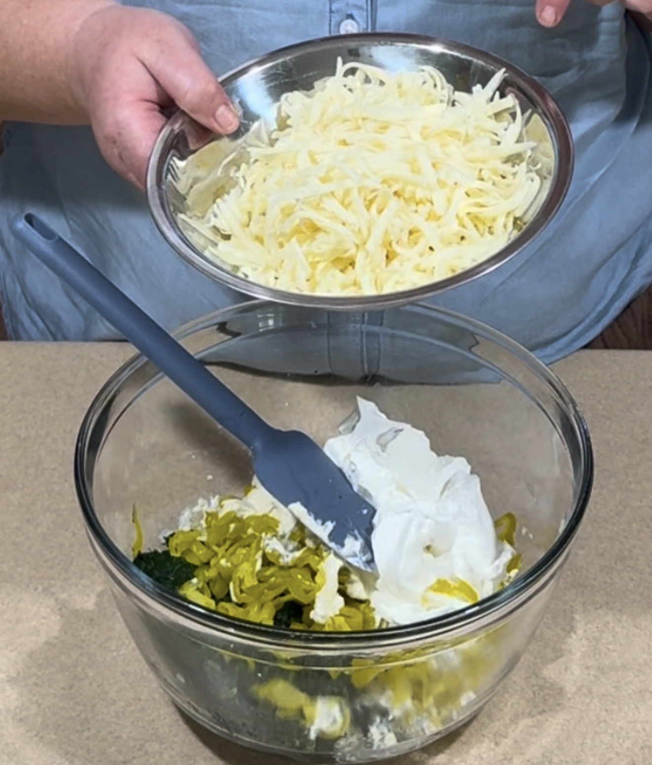 A person holds a plate of shredded cheese above a bowl containing sour cream, chopped vegetables, and a gray spatula, preparing to combine the ingredients.