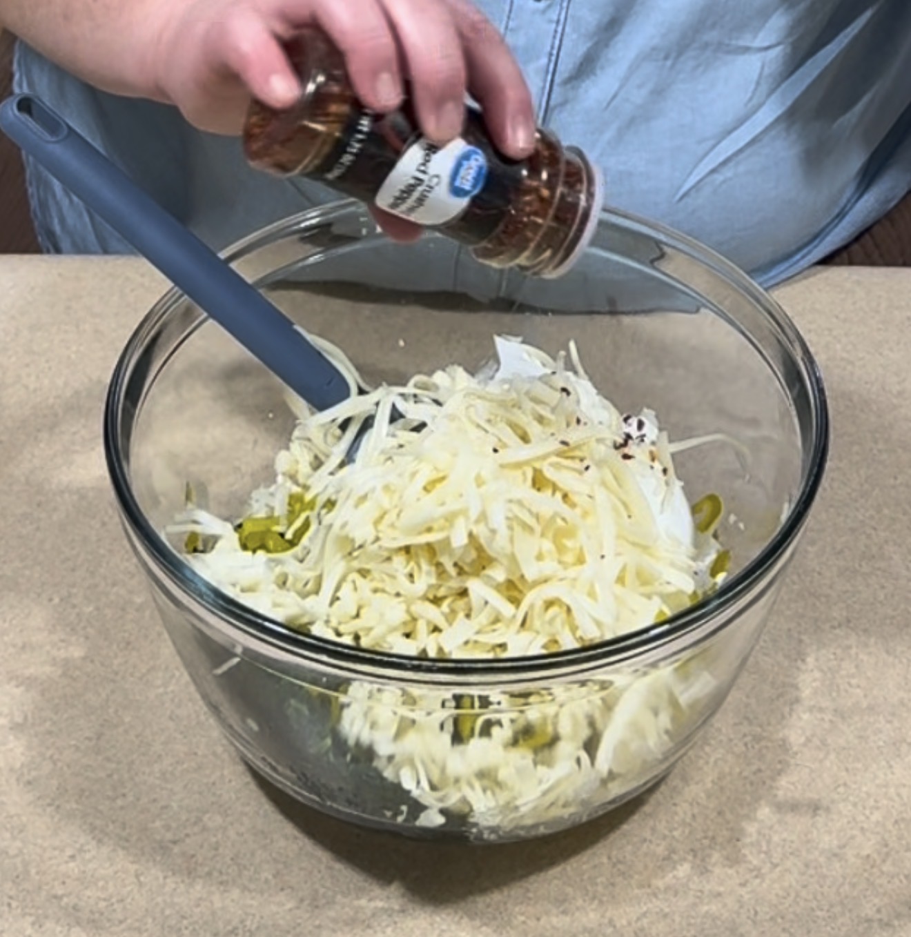 A person sprinkles red pepper flakes from a jar into a glass bowl filled with shredded cheese, sliced jalapeños, and other ingredients. A gray spatula rests in the bowl.