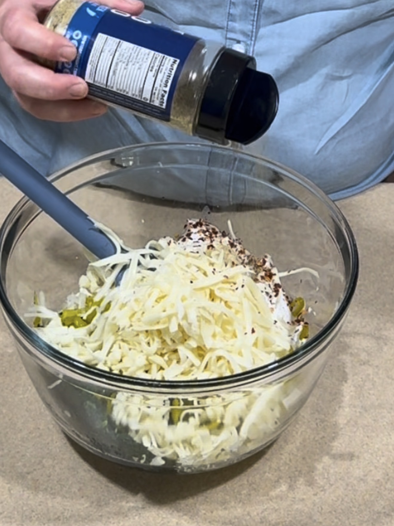 A person sprinkles seasoning from a bottle into a glass bowl filled with shredded cheese, cream cheese, and chopped green ingredients, while holding a blue spatula.