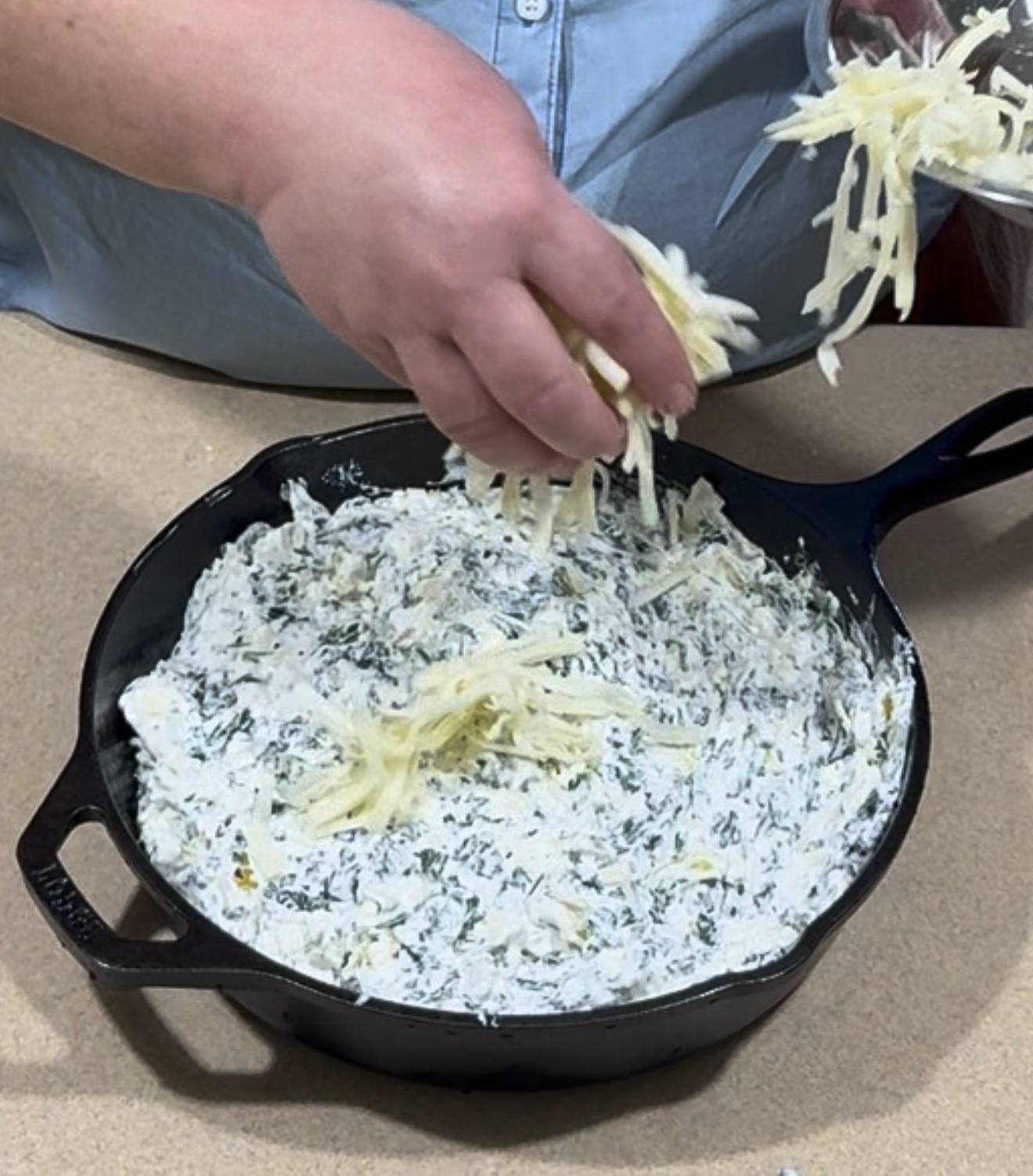 A person sprinkles shredded cheese onto a creamy spinach mixture in a black cast iron skillet, preparing a dish on a tan countertop.