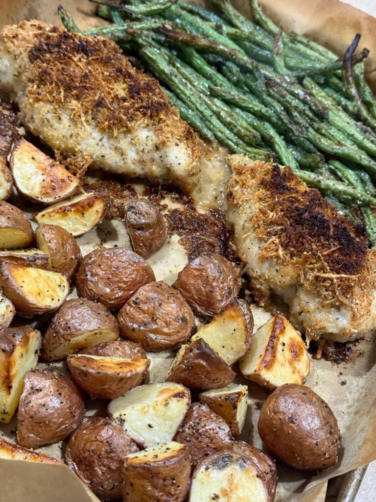 Sheet pan with roasted red potatoes, breaded chicken breasts, and green beans on parchment paper. The food looks golden brown and well-seasoned, with a crispy coating on the chicken and roasted vegetables.