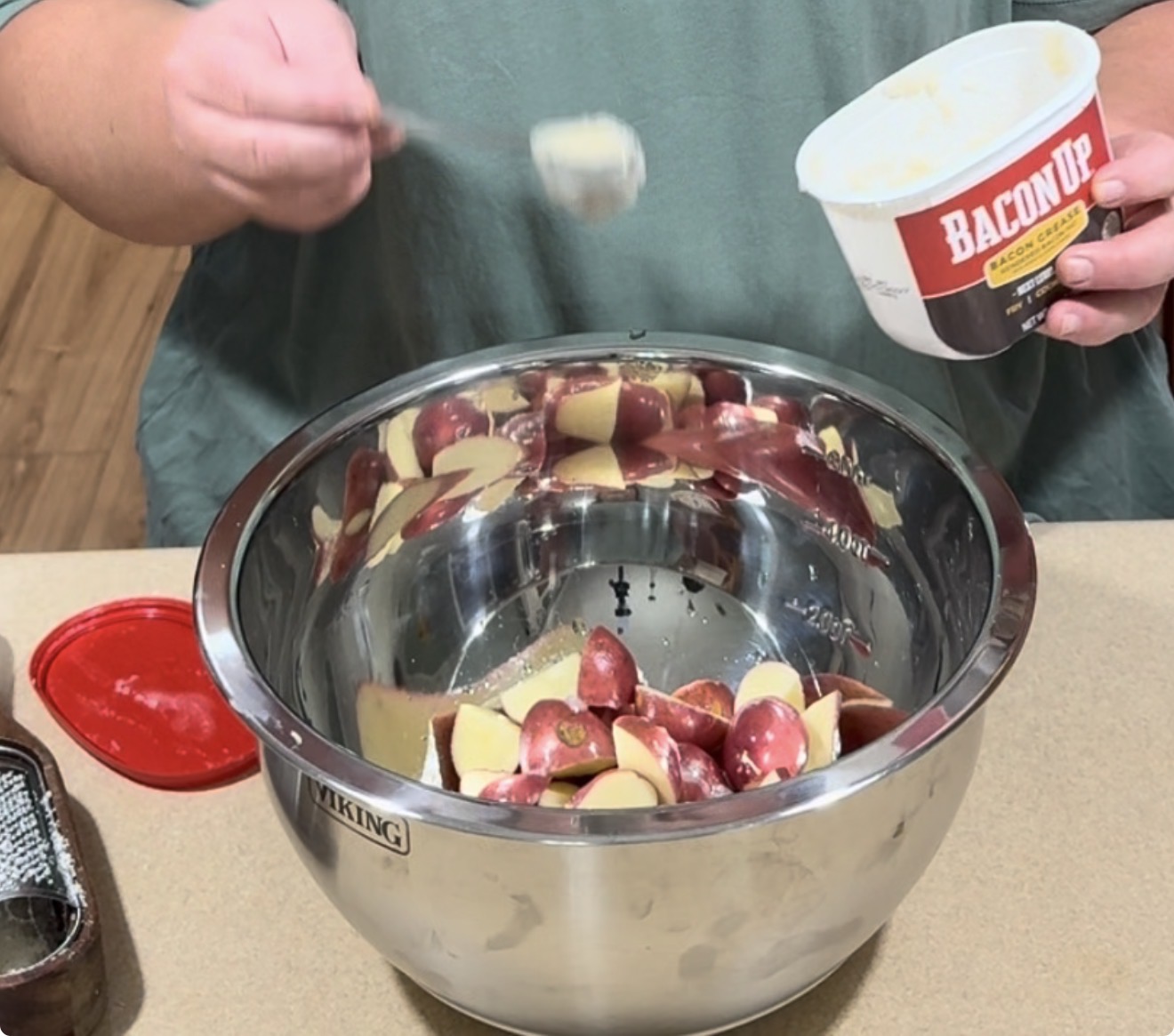 Person scooping Bacon Up bacon grease from a tub into a metal bowl filled with sliced red potatoes on a kitchen counter.