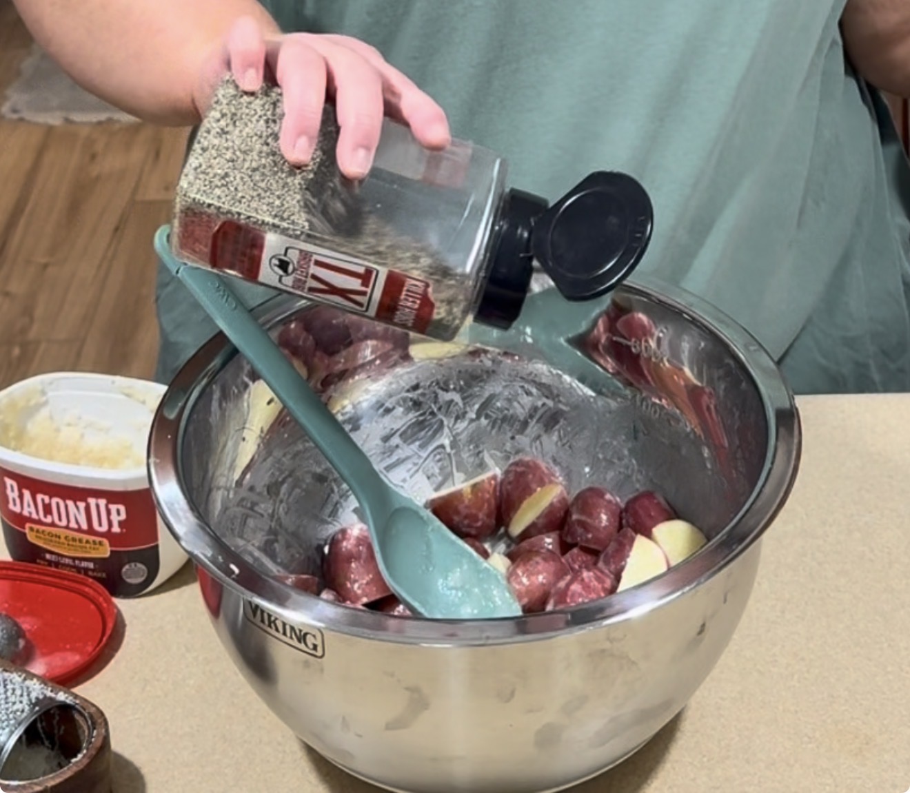 A person sprinkles black pepper on halved red potatoes in a large metal mixing bowl, using a green spatula to mix. A container labeled Bacon Up is nearby on the countertop.