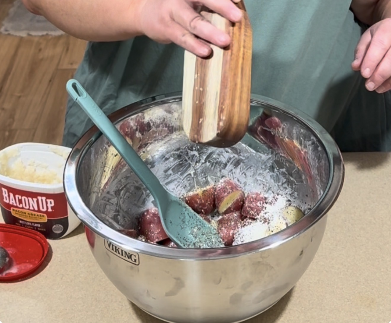 A person seasons cut red potatoes in a metal mixing bowl with a wooden shaker. A blue spatula rests in the bowl. A tub labeled Bacon Up sits on the counter nearby.