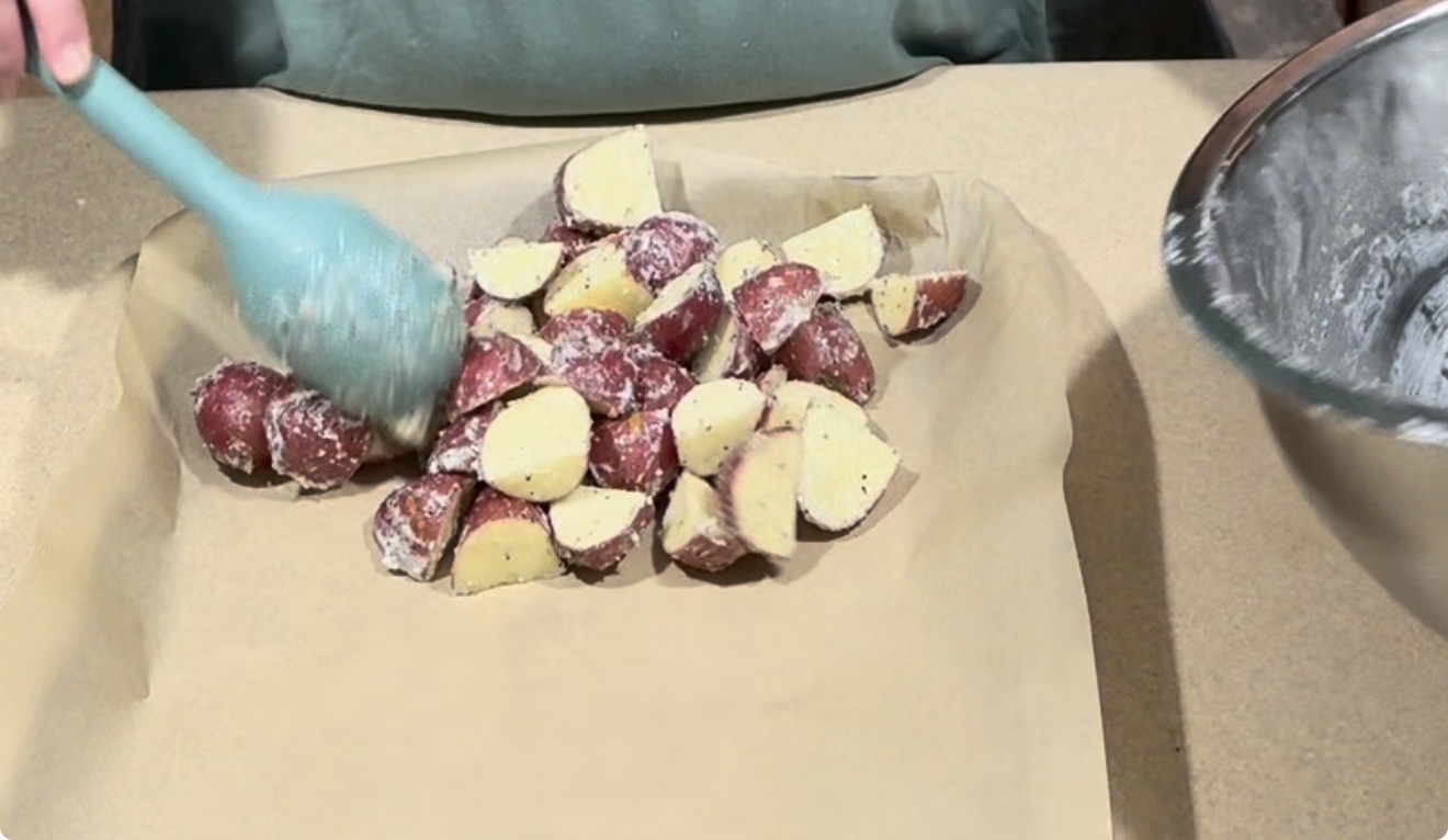 Red potatoes coated in seasoning are spread on a parchment-lined baking sheet with a blue spatula; a mixing bowl with residue is visible on the right side.