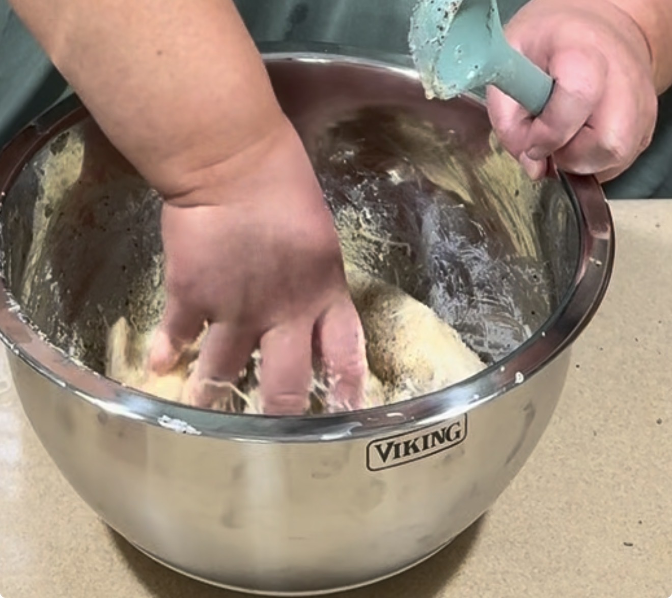 A person mixing dough by hand in a large stainless steel bowl labeled Viking on a countertop, holding a blue spatula in the other hand.