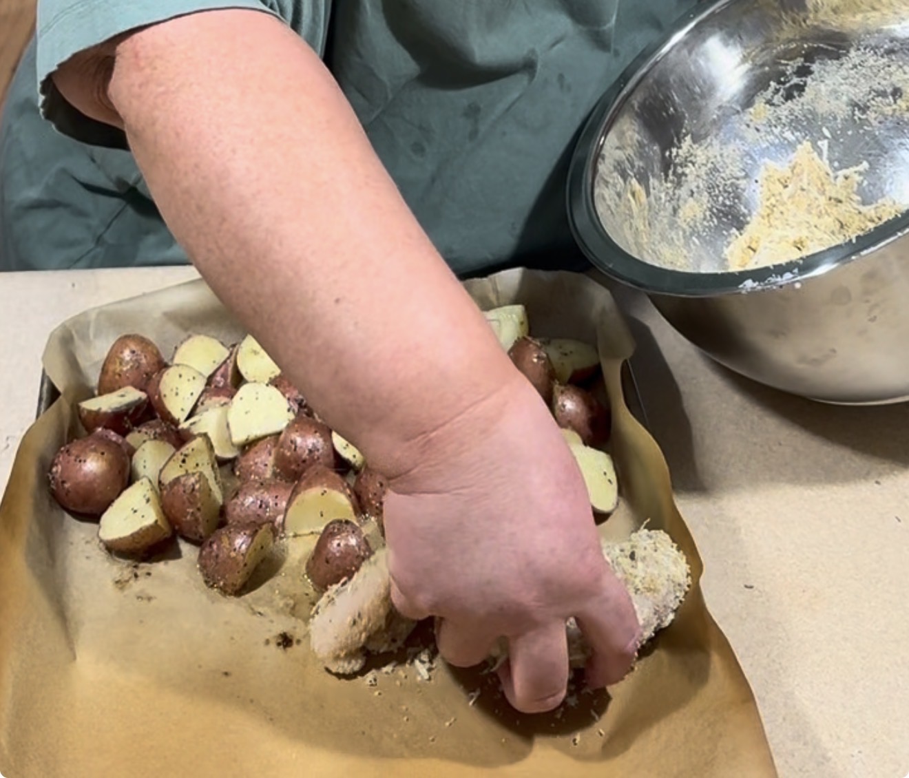 A person coats a chicken drumstick in breadcrumbs over a tray of seasoned, halved red potatoes. A metal bowl with more breadcrumb mixture sits nearby on a parchment-lined baking sheet.