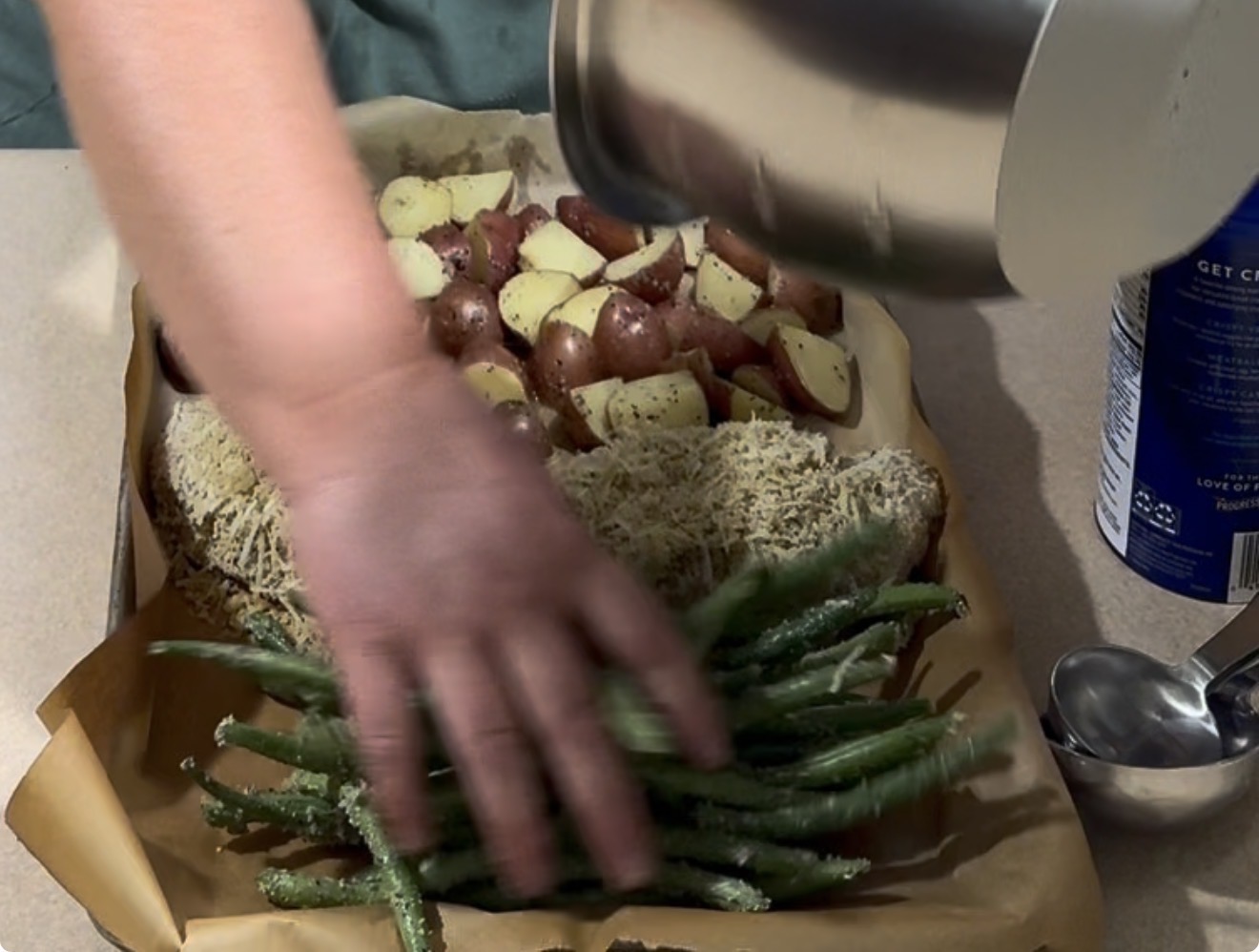 A hand arranges green beans on a baking tray with seasoned potatoes and a cheese-topped entrée, while another hand pours something from a metal bowl. A canister of salt and metal spoons are nearby.