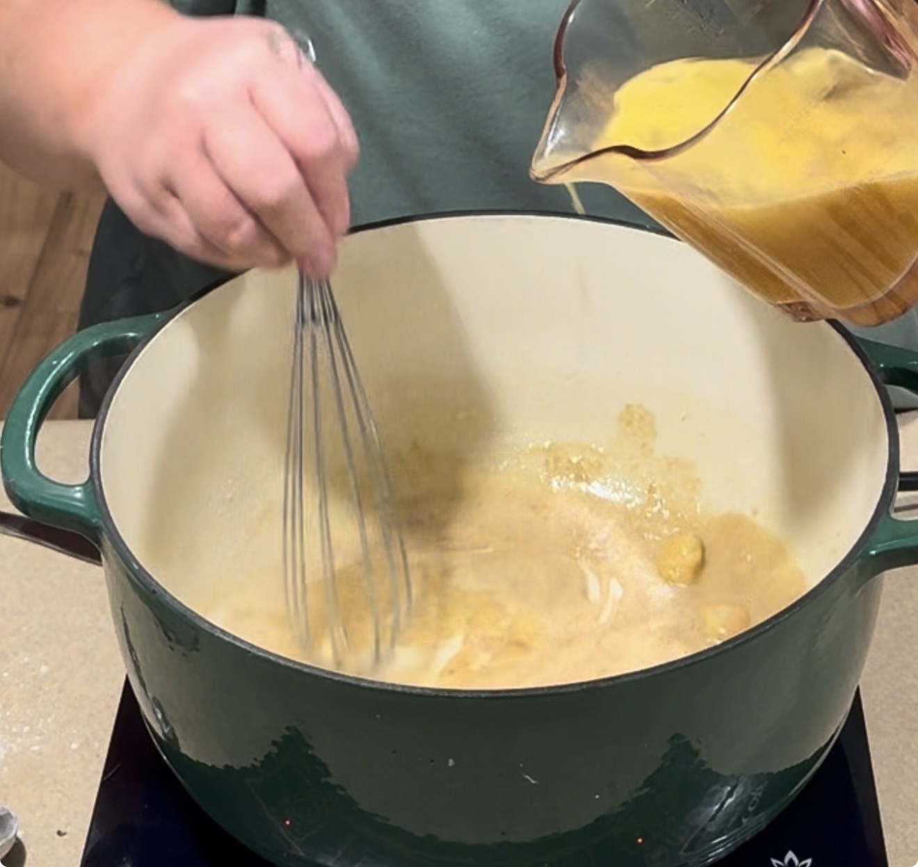 A person whisks a roux in a green Dutch oven while pouring a yellow liquid from a measuring cup into the pot on a stovetop.