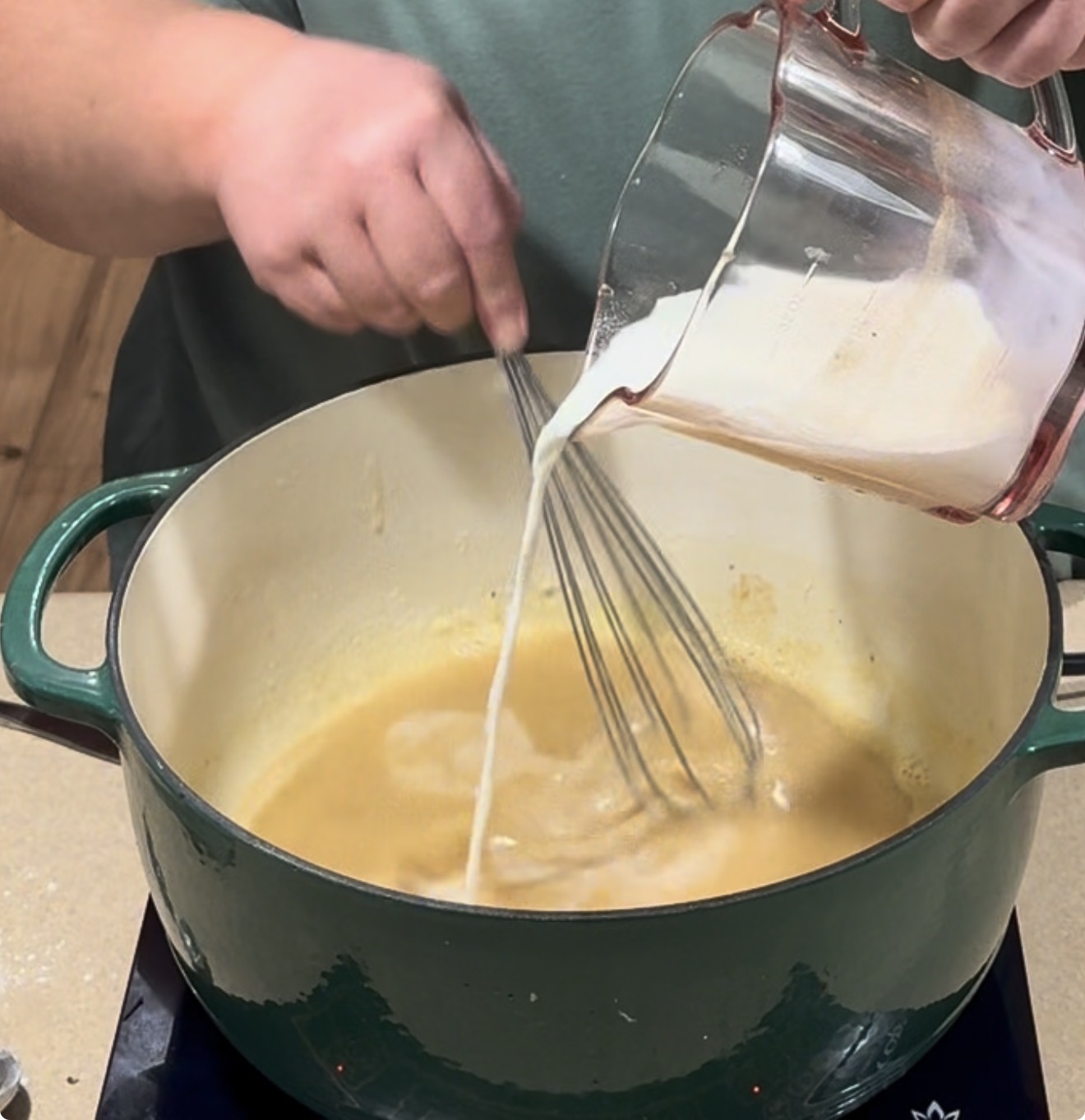 A person whisks a creamy liquid into a yellow mixture in a large green Dutch oven on a stovetop. The liquid is being poured from a glass measuring cup.