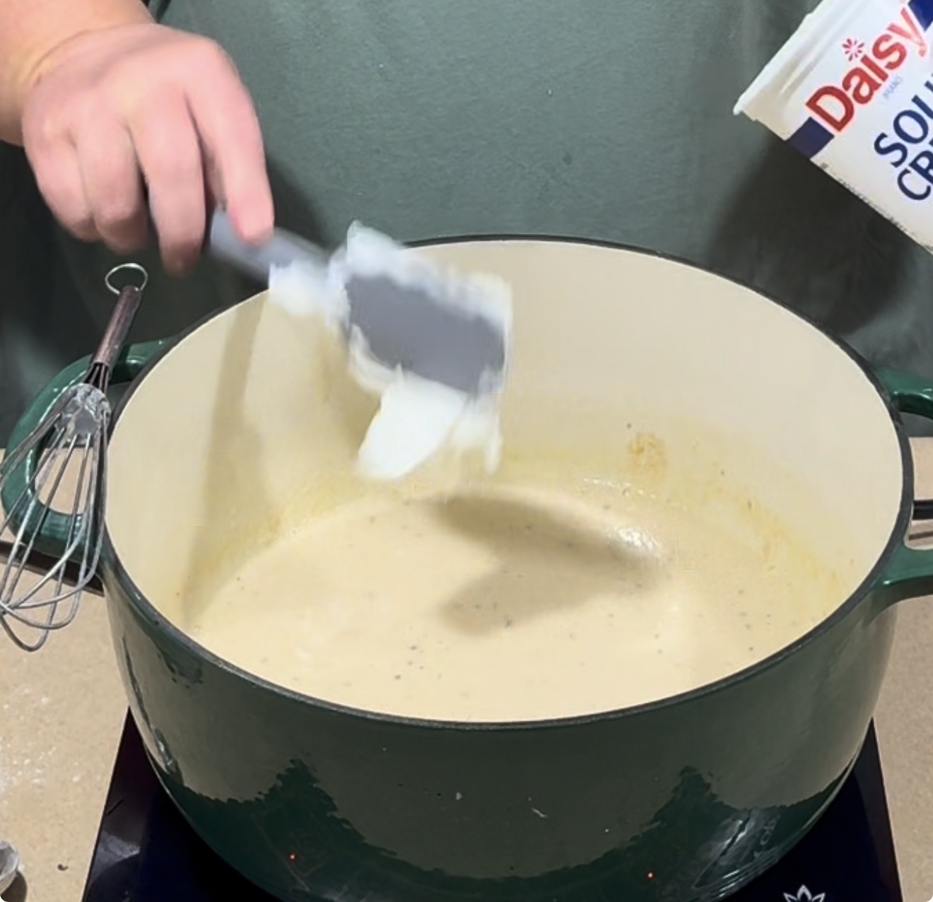 A person adds sour cream from a container into a green Dutch oven filled with a creamy mixture on a stovetop. A whisk rests nearby.
