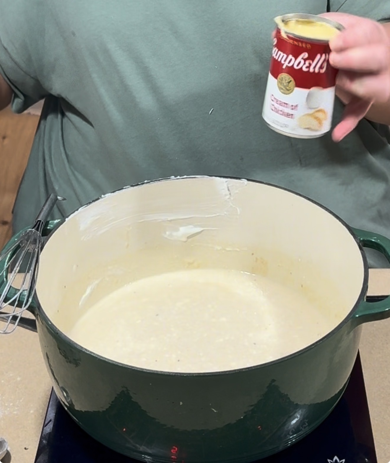 A person in a green shirt holds a can of Campbells cream of chicken soup above a green Dutch oven filled with a creamy mixture, with a whisk nearby.