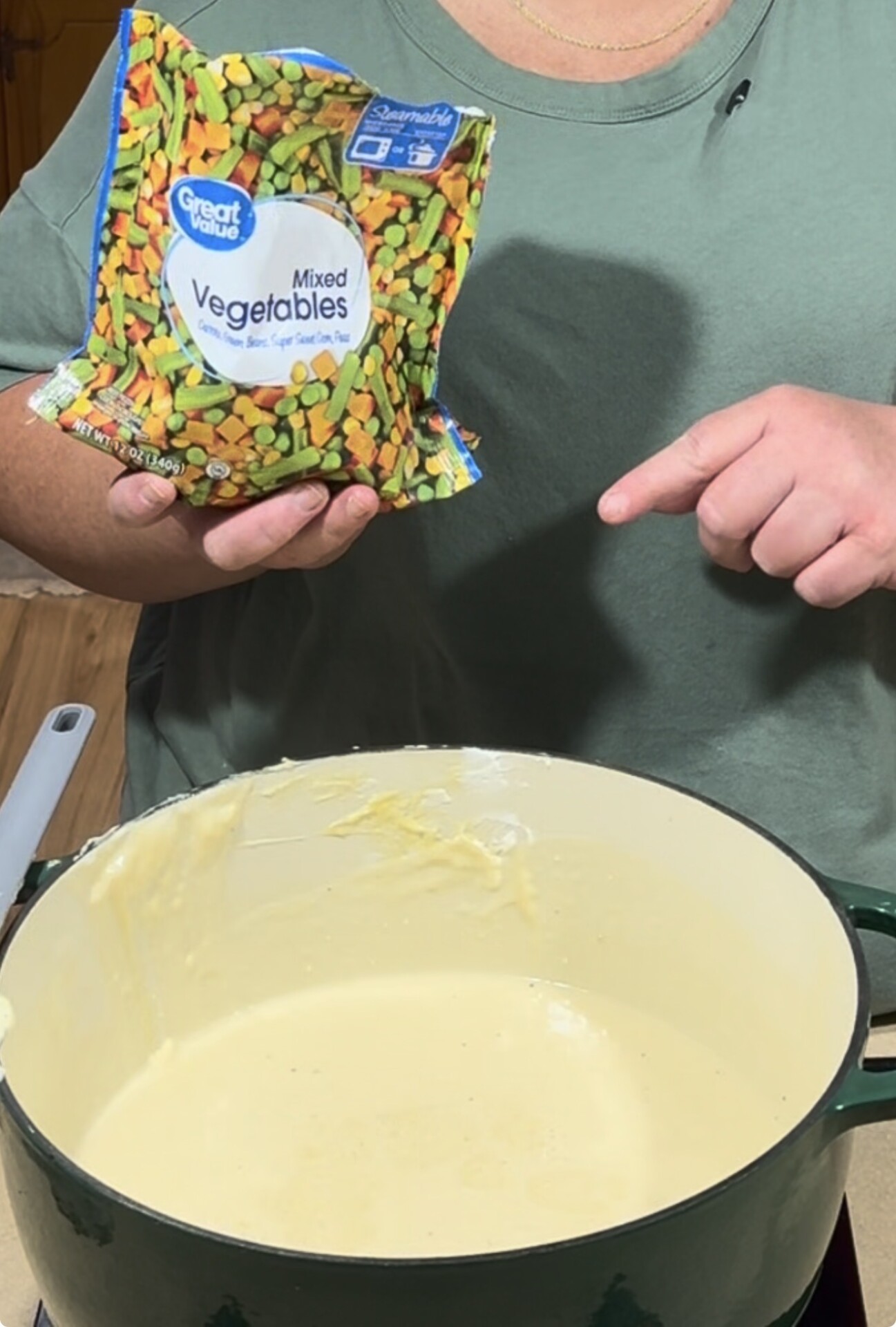 A person holds a bag of frozen mixed vegetables and points to a pot filled with a creamy mixture on a stove.