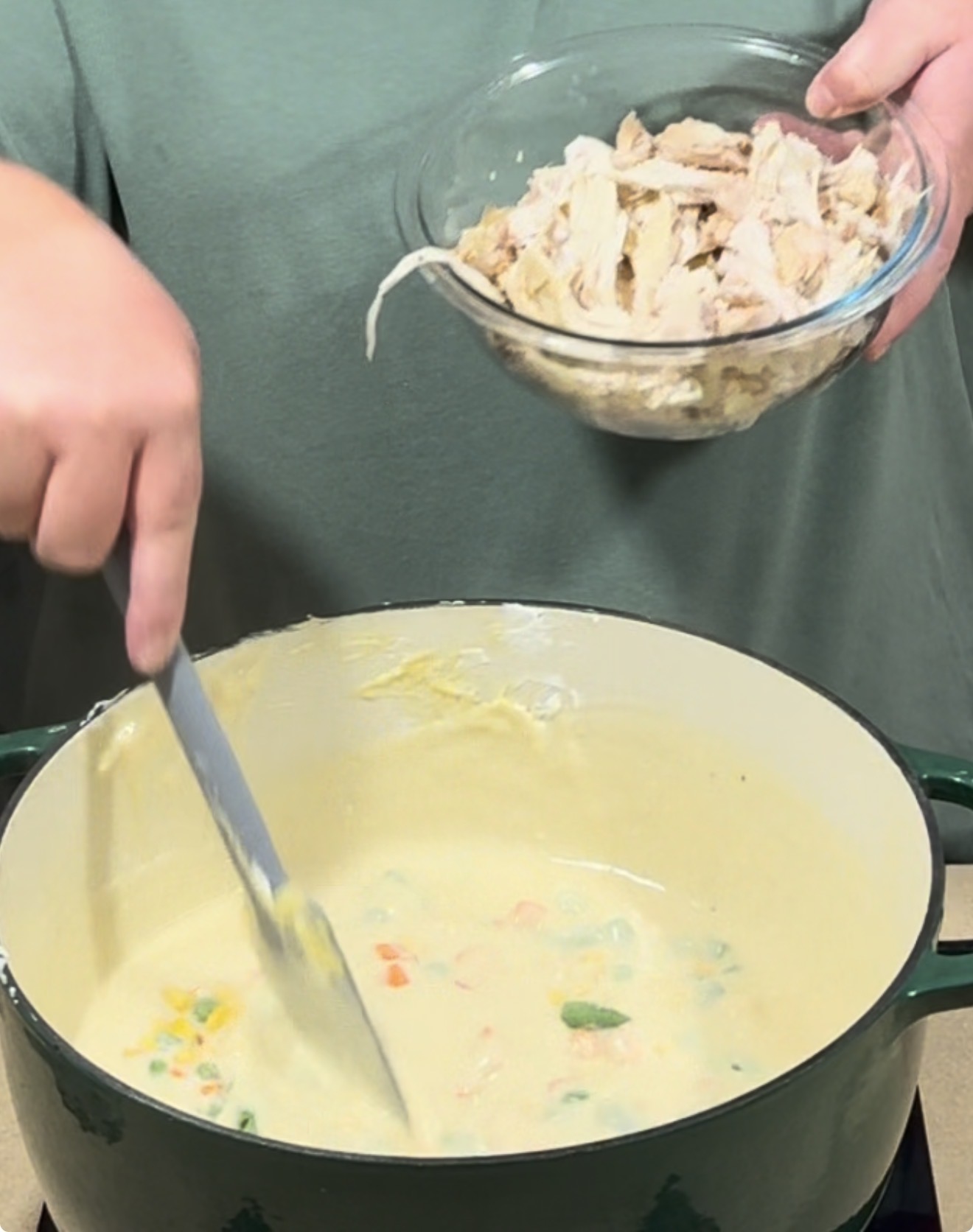 A person holding a bowl of shredded chicken while stirring a pot of creamy vegetable soup on a stovetop.