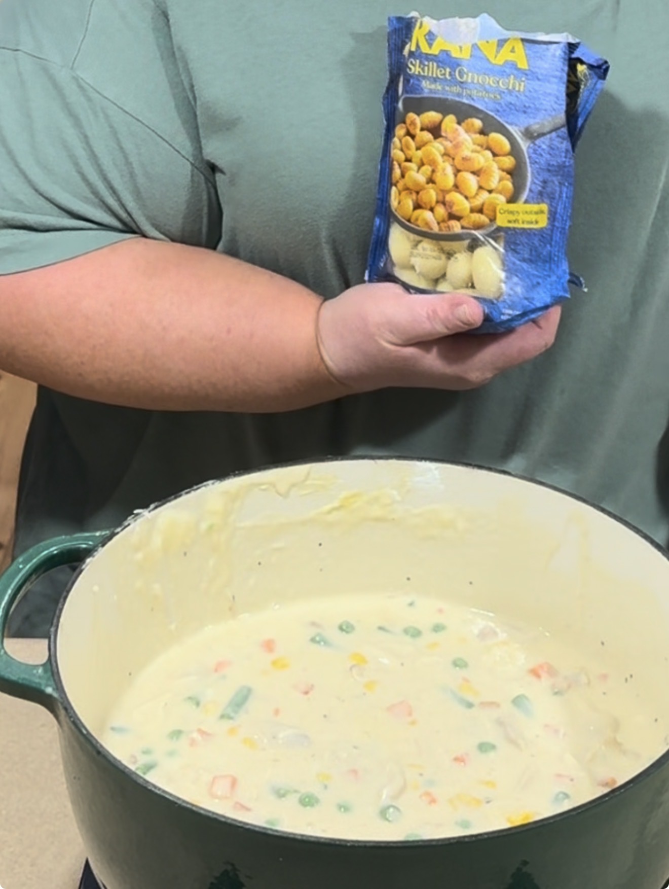 A person holds a bag of skillet gnocchi above a green pot filled with creamy soup containing mixed vegetables.