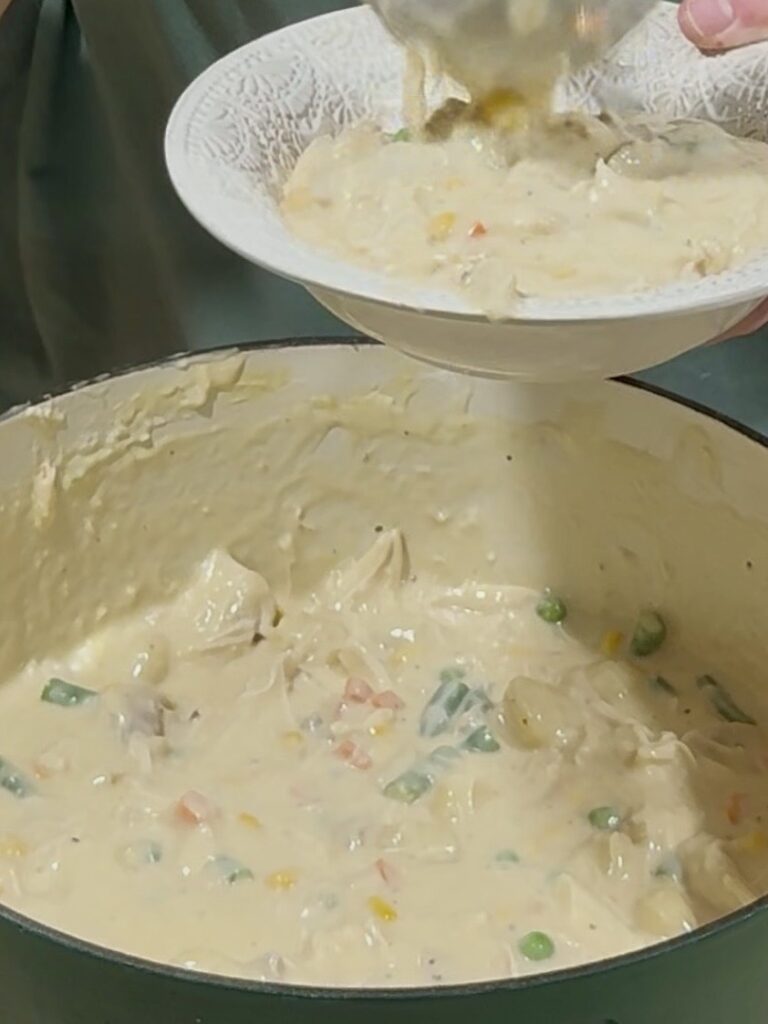 A person serves creamy chicken and vegetable stew with peas, carrots, and corn from a large pot into a white bowl.