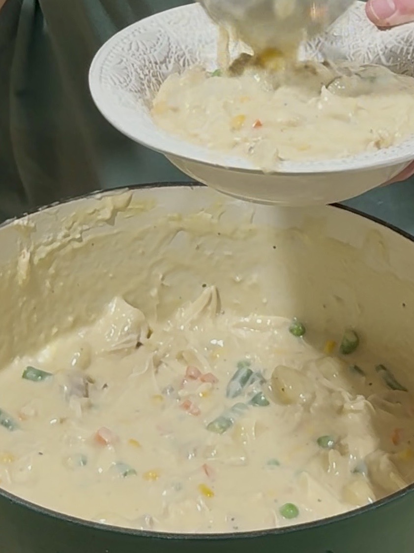 A person serves creamy chicken and vegetable stew with peas, carrots, and corn from a large pot into a white bowl.