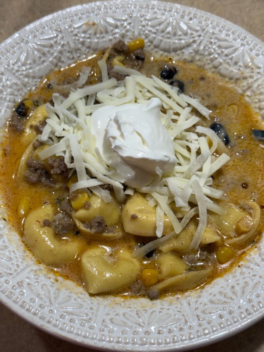 A bowl of soup with tortellini, ground meat, corn, black beans, shredded cheese, and a dollop of sour cream on top, served in a white textured bowl.