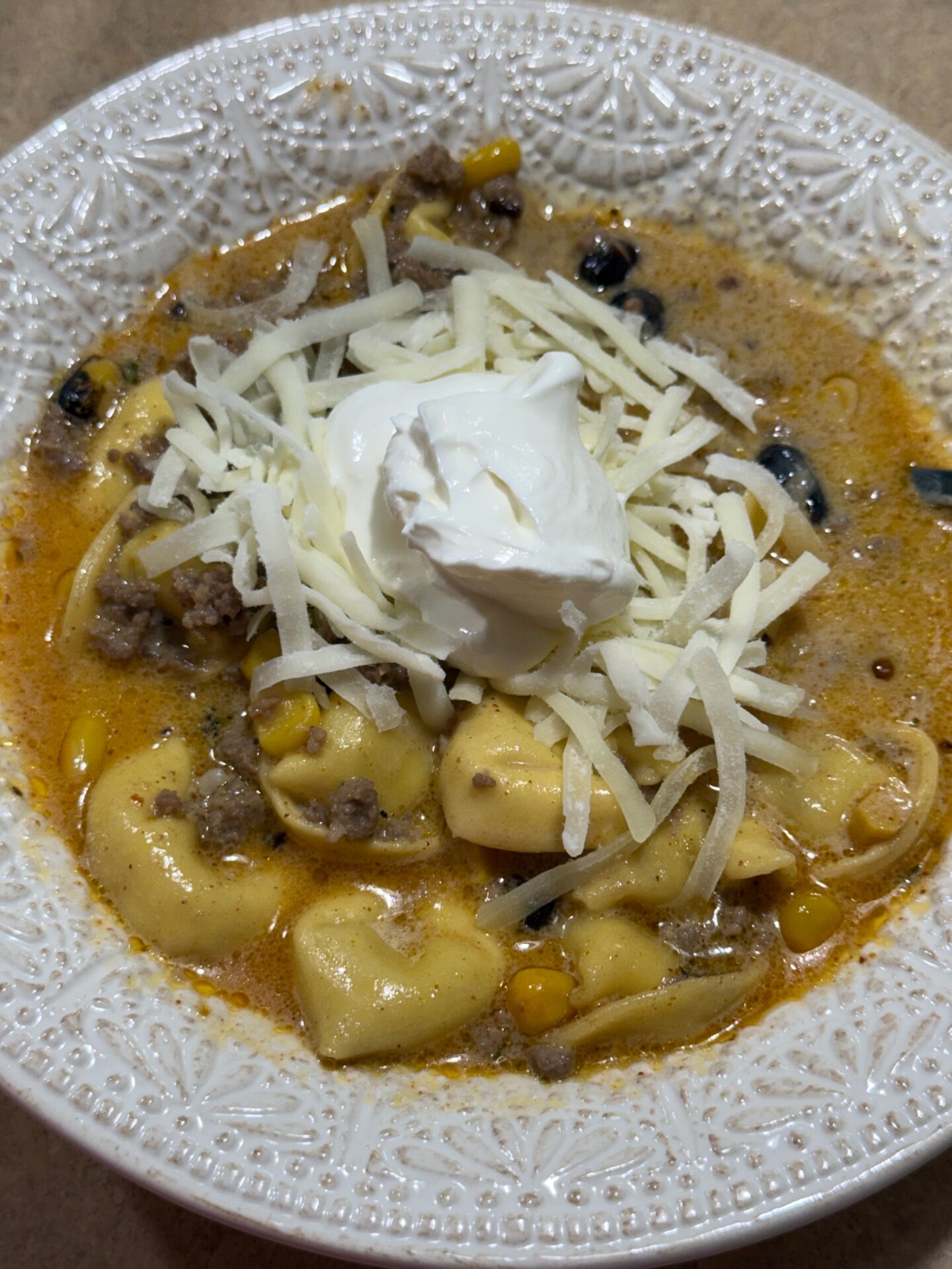 A bowl of soup with tortellini, ground meat, corn, black beans, shredded cheese, and a dollop of sour cream on top, served in a white textured bowl.