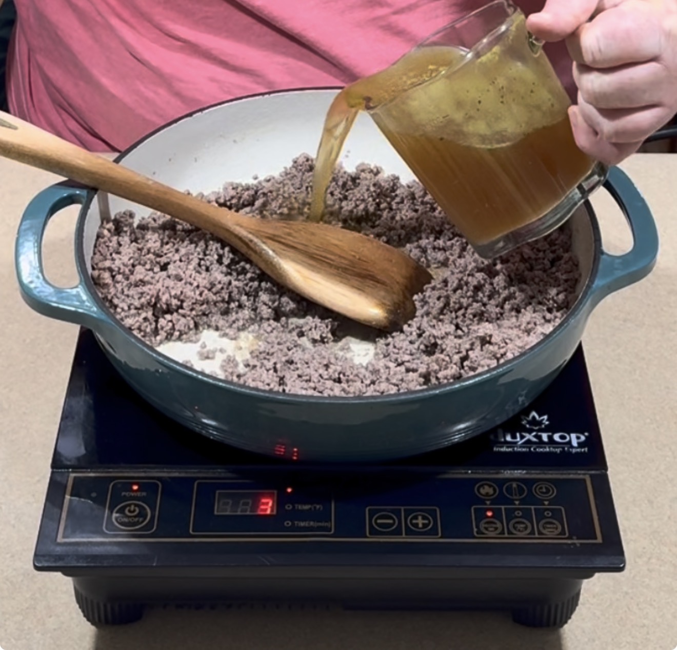 A person pours broth from a glass pitcher into a pot of cooked ground beef, stirring with a wooden spoon on an induction cooktop.