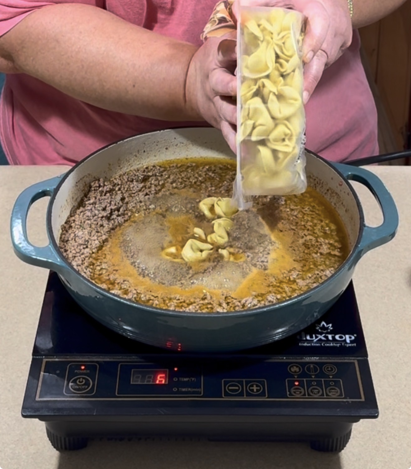 A person pours cheese tortellini into a pot of browned ground meat and broth on an electric stove.