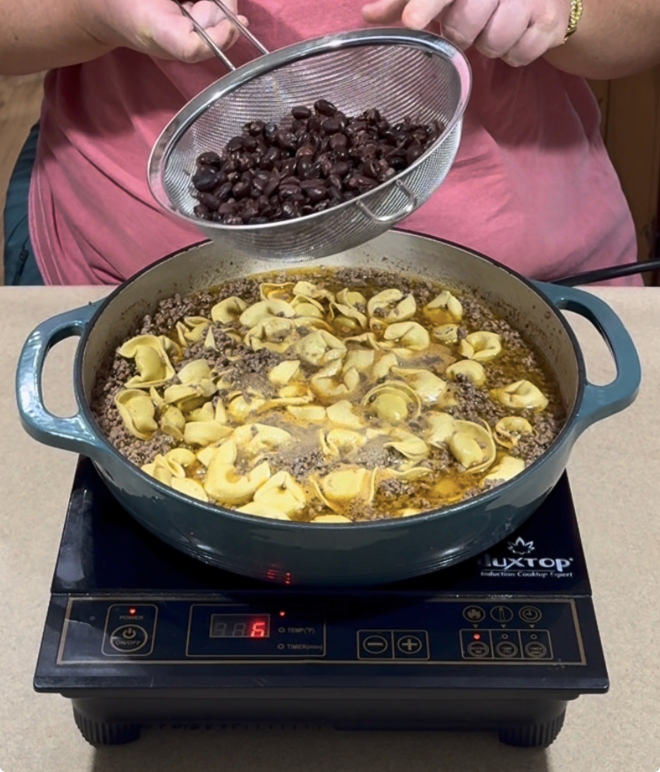 A person holds a strainer of black beans over a pot on an induction cooktop. The pot contains sliced mushrooms, ground meat, and broth.