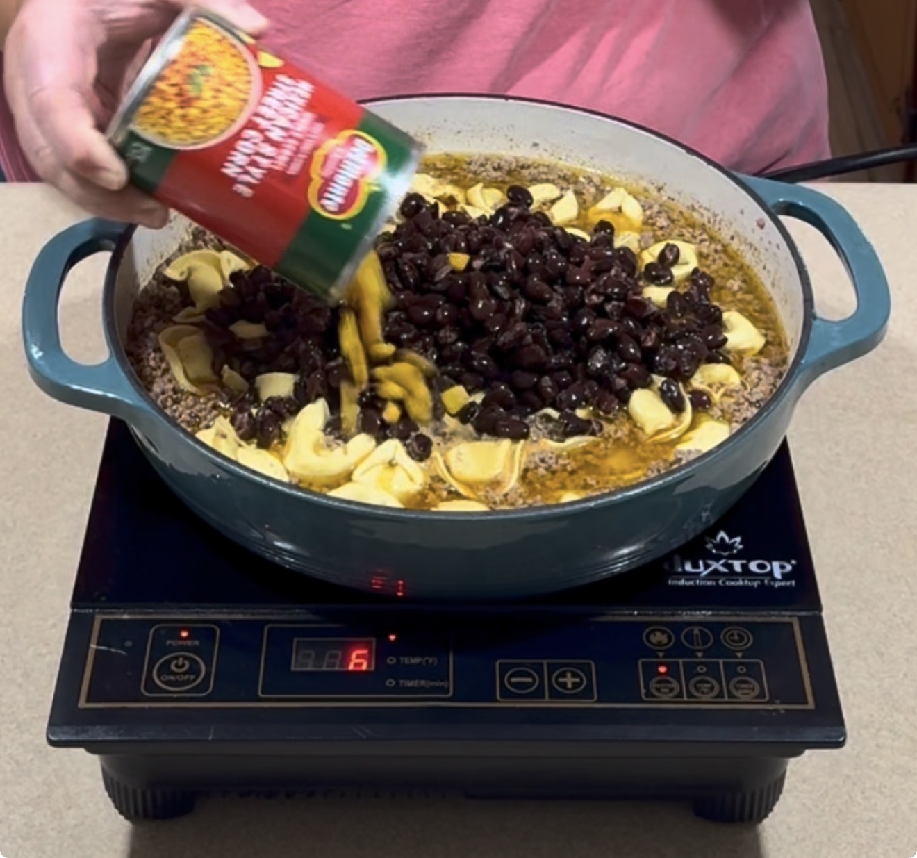 A person pours a can of mixed vegetables into a pot already containing black beans and sliced yellow vegetables on an induction cooktop.