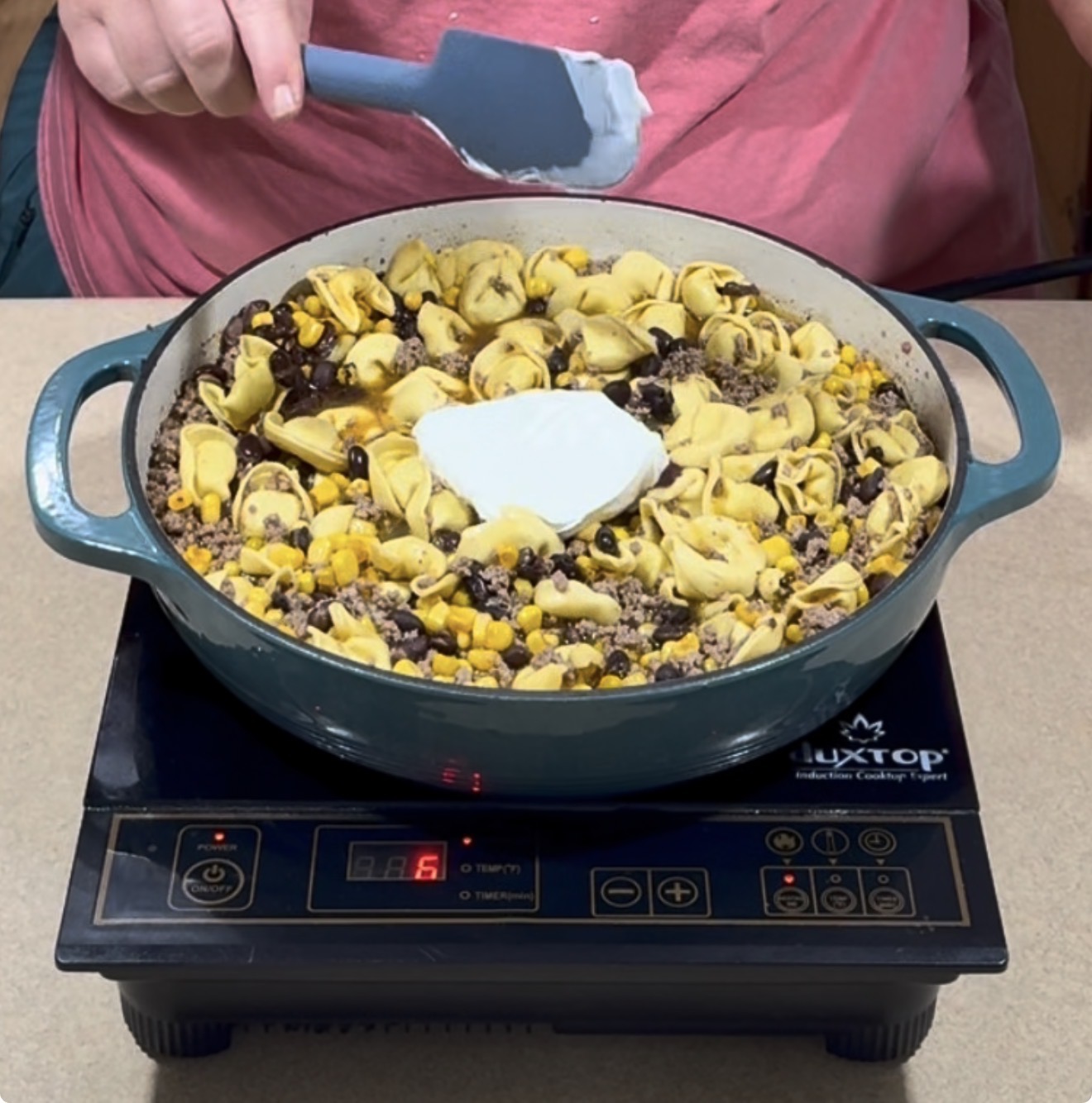 A person in a pink shirt stirs a skillet of cooked tortellini, ground beef, black beans, corn, and a dollop of sour cream on a portable stovetop.