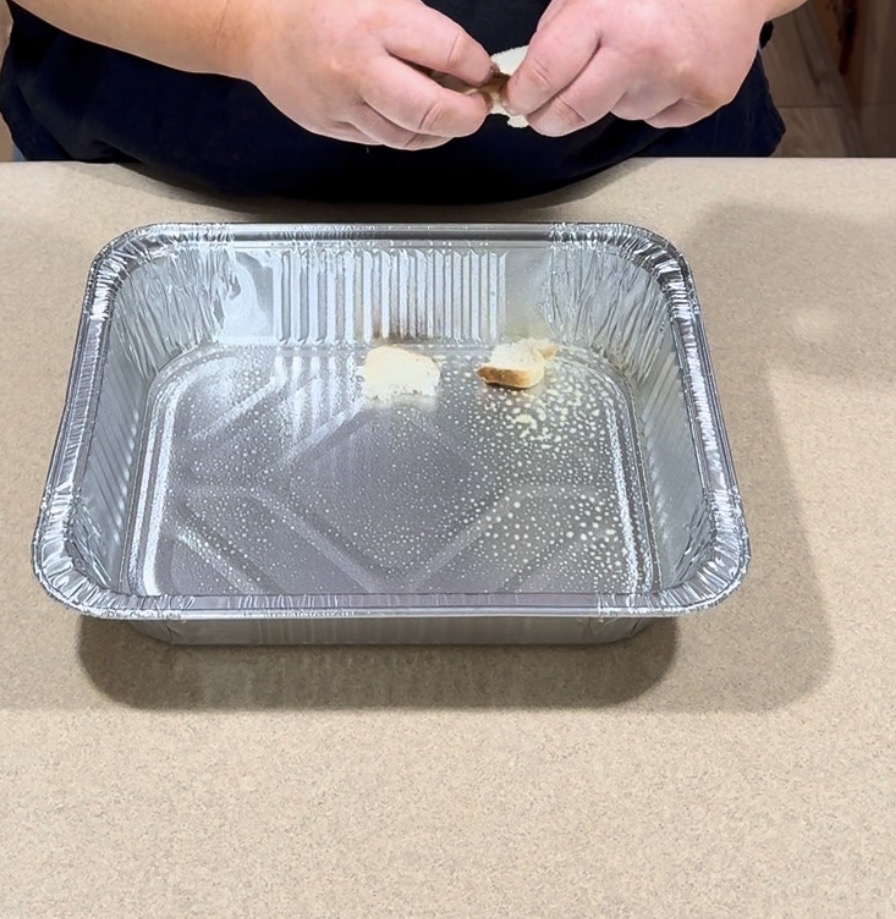 A persons hands are breaking bread rolls over a large, empty aluminum baking pan on a beige countertop.