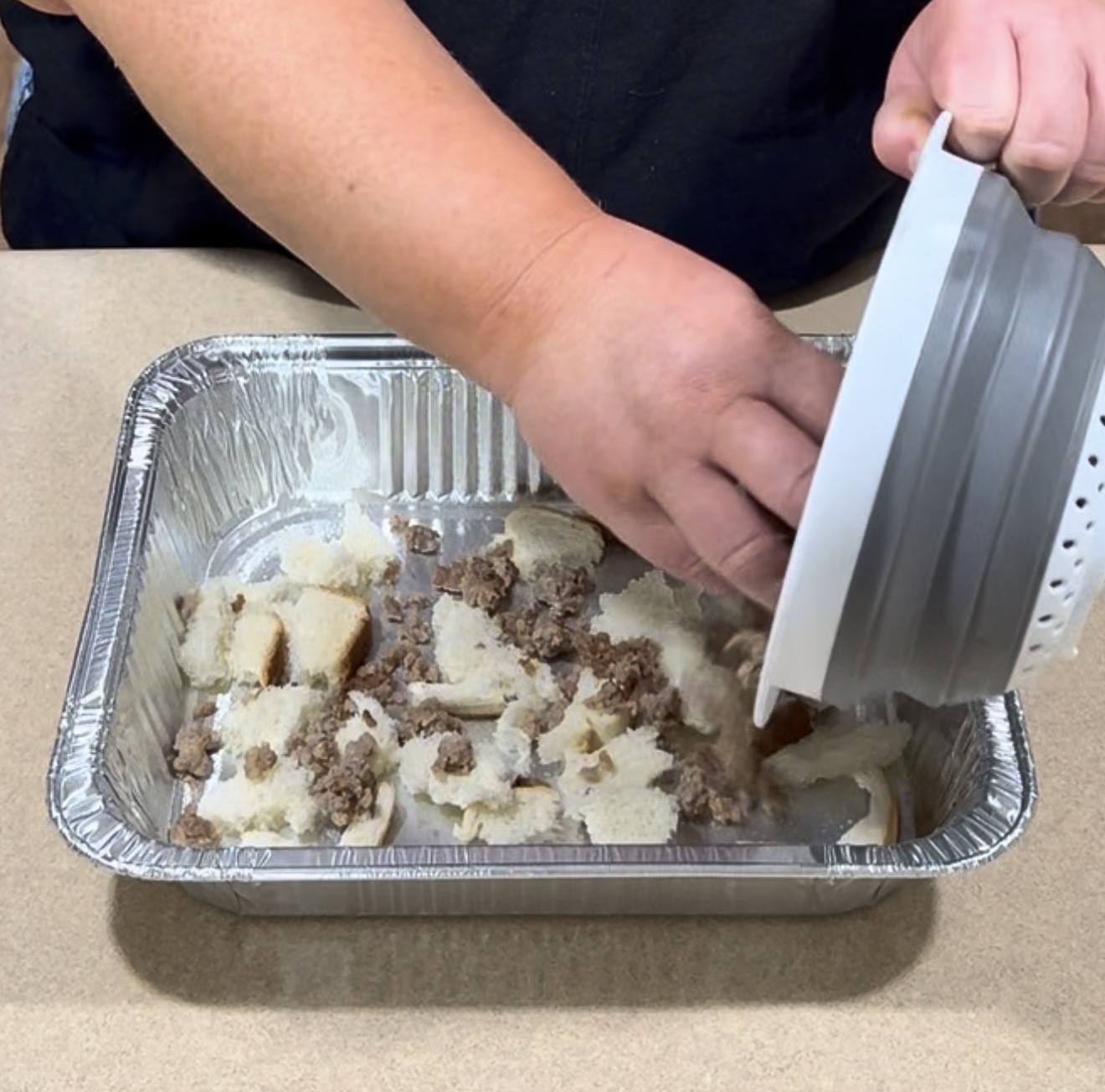 A person adds ground meat and torn bread pieces from a bowl into a rectangular aluminum tray on a countertop.