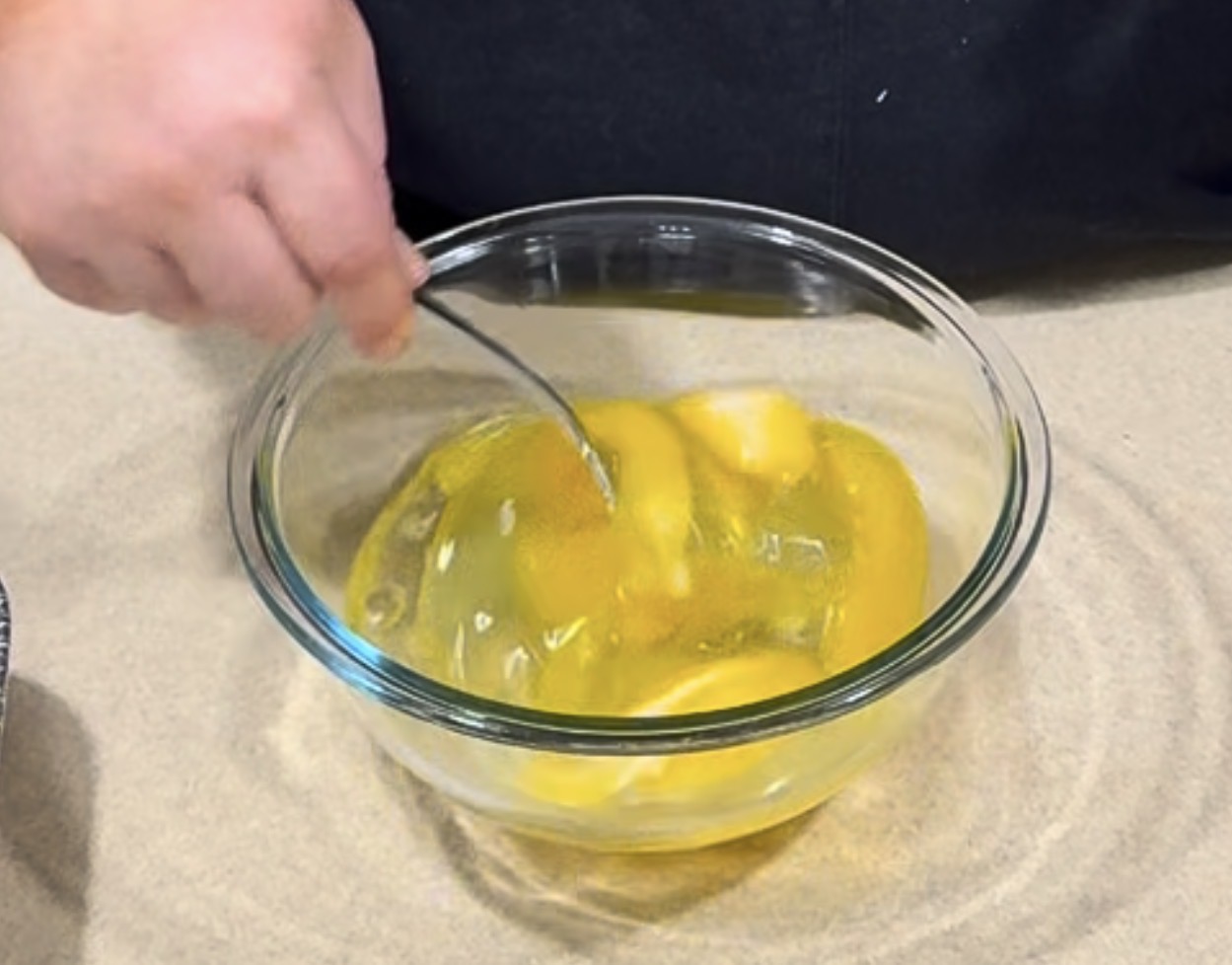 A person uses a fork to whisk raw eggs in a transparent glass bowl on a countertop.