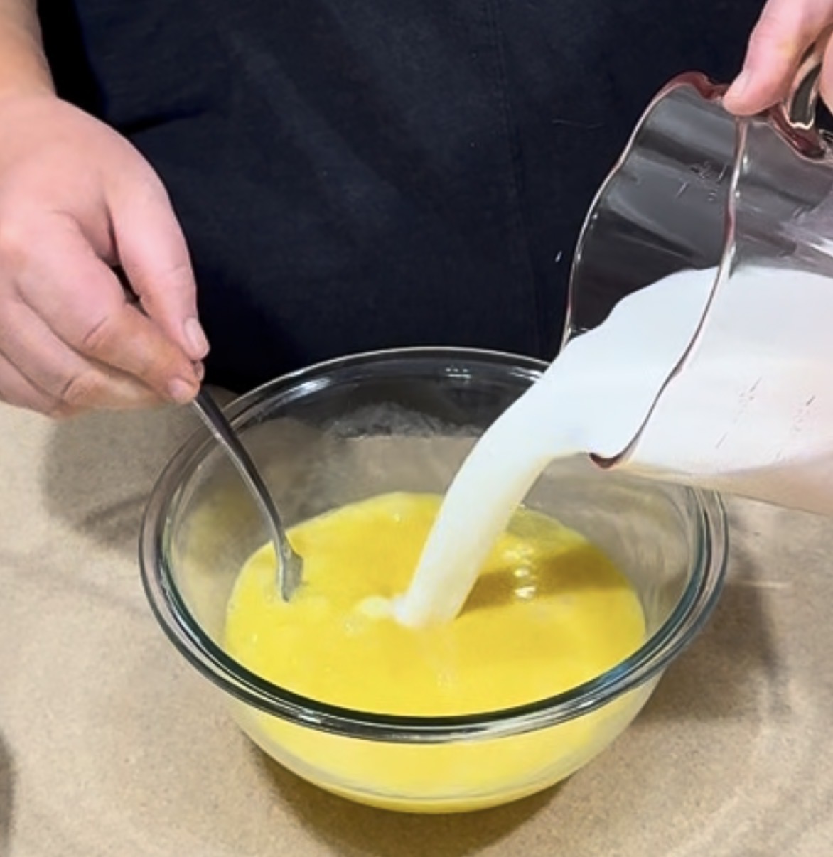 A person pours milk from a measuring cup into a glass bowl with beaten eggs while stirring with a spoon.