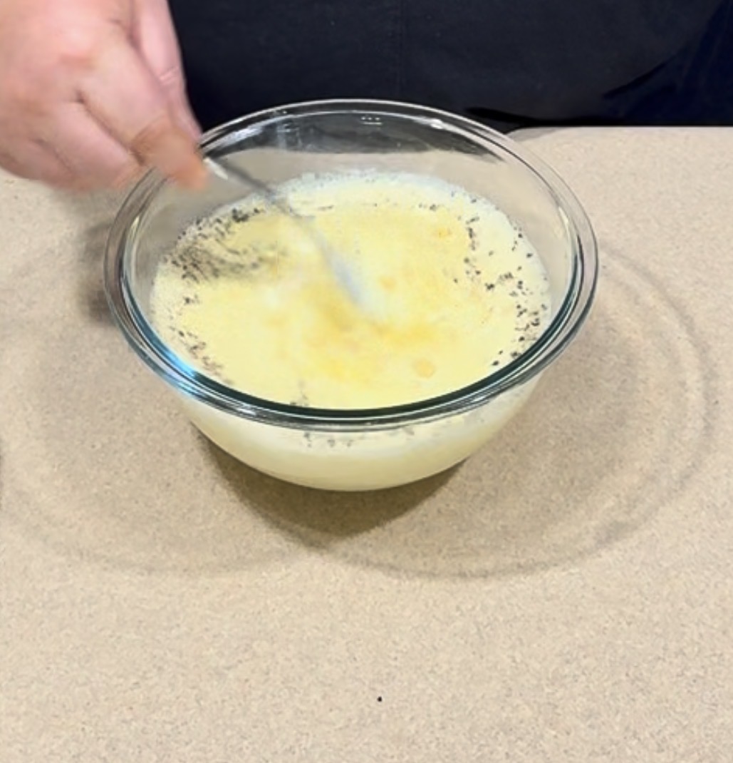 A person stirs a yellow liquid mixture in a clear glass bowl with a metal spoon on a beige countertop.