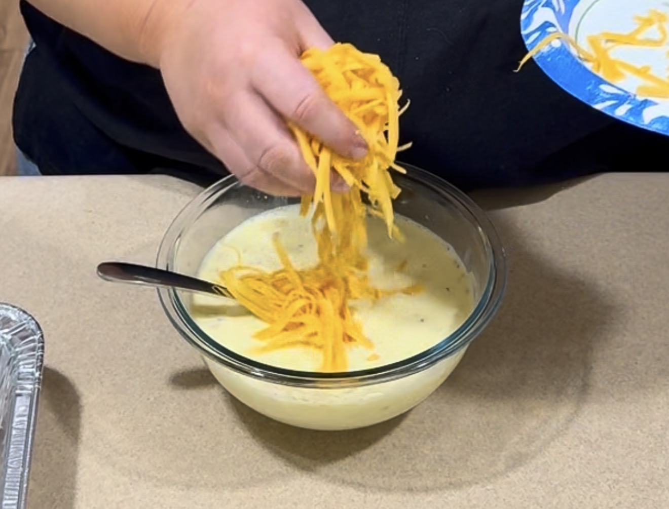 A person adds shredded cheddar cheese to a bowl of creamy mixture with a spoon in it, preparing food on a countertop.