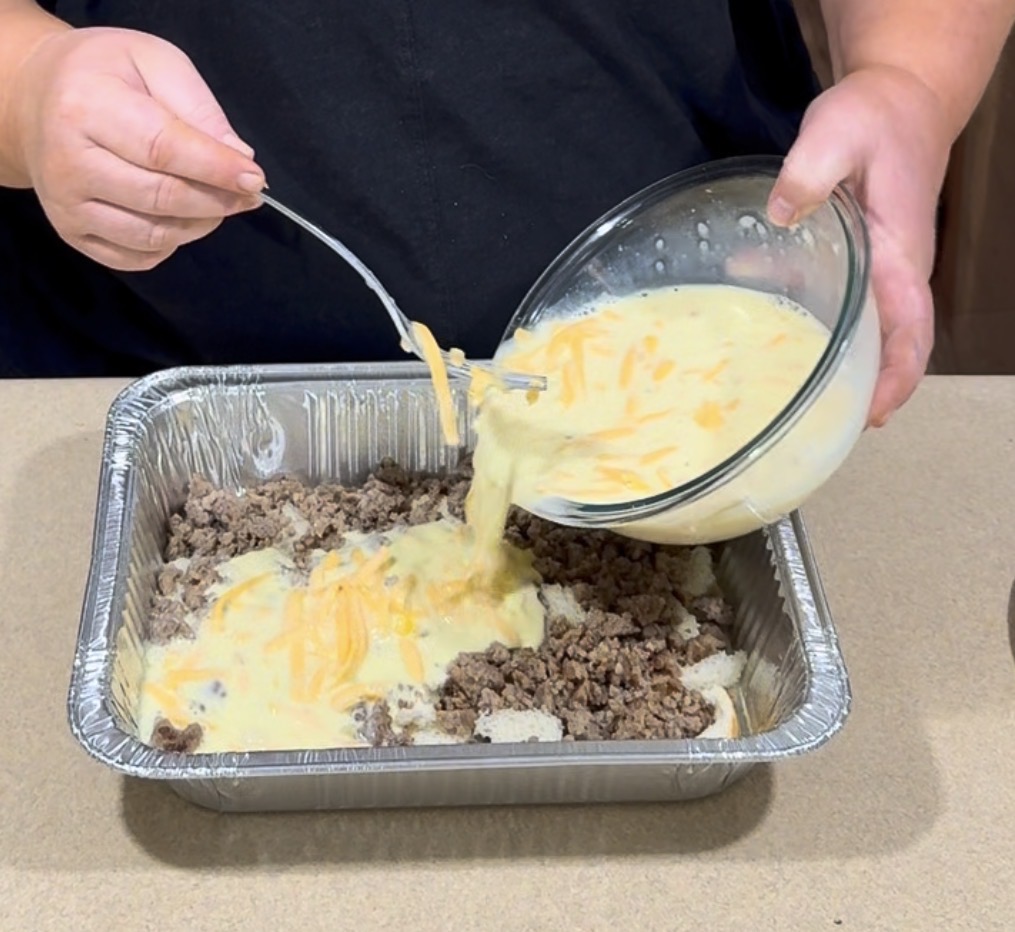 A person pours a mixture of eggs and shredded cheese from a glass bowl onto cooked ground beef in a foil tray, preparing a casserole.