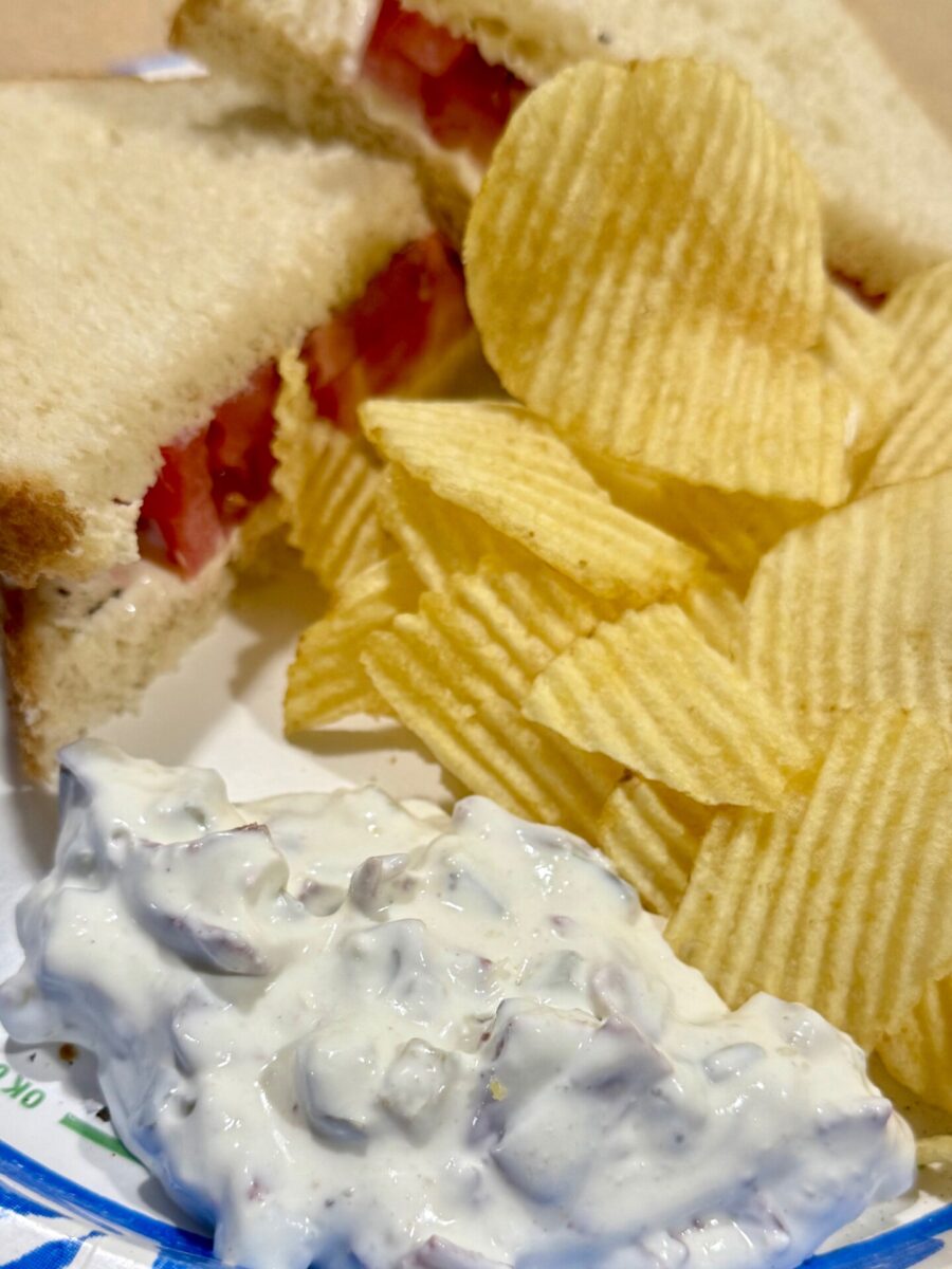 A plate with a tomato sandwich on white bread, a pile of ridged potato chips, and a serving of creamy dip with visible chunks.