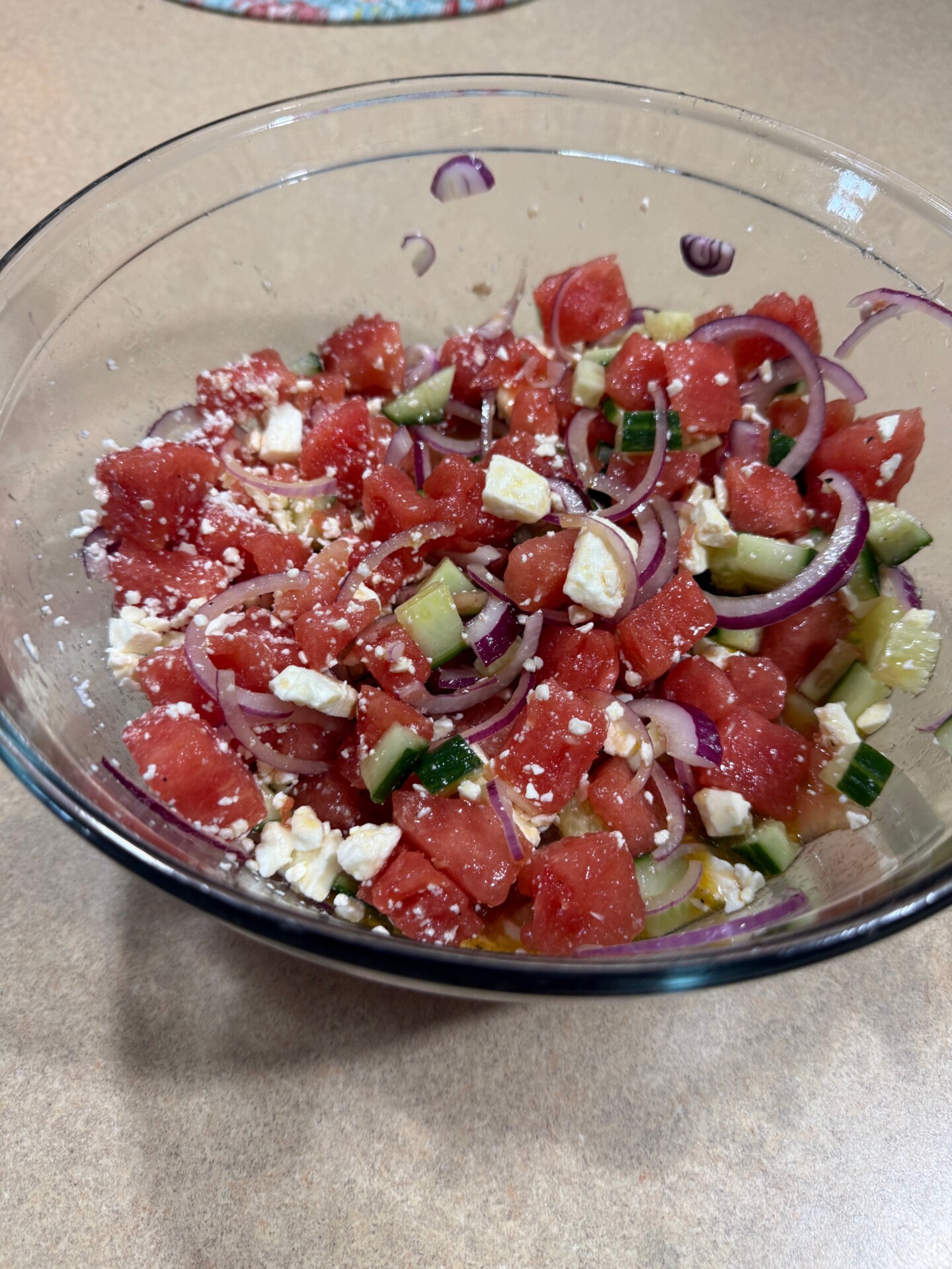 A glass bowl filled with a salad of diced watermelon, cucumber, red onion slices, and crumbled feta cheese, sitting on a beige countertop.