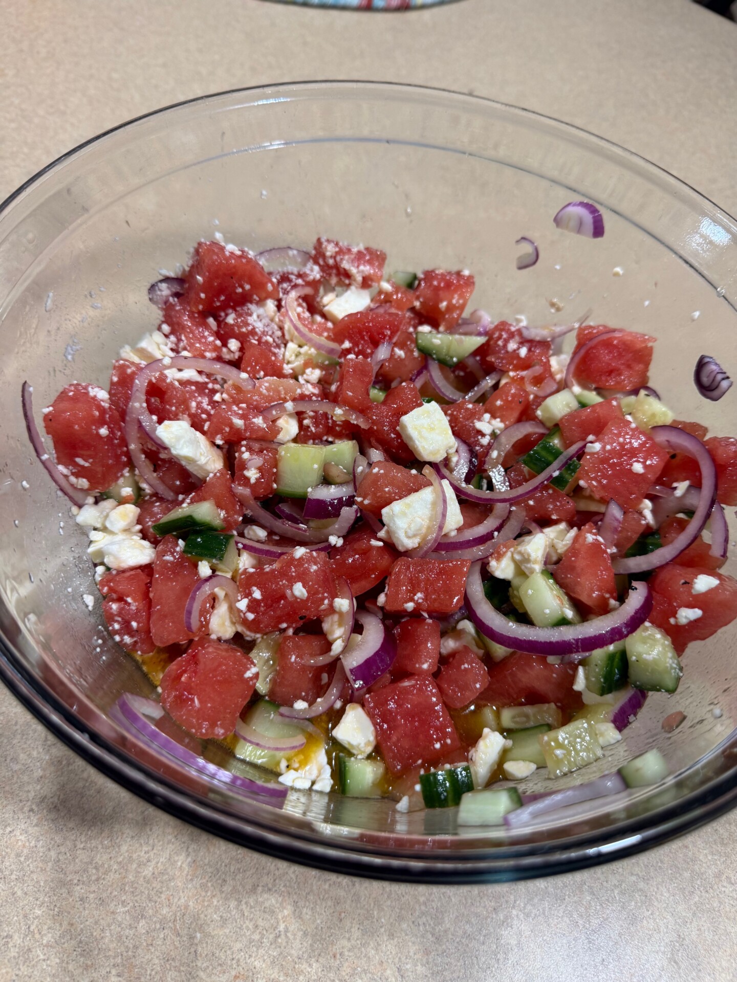 A glass bowl contains a salad of watermelon cubes, cucumber pieces, red onion slices, crumbled feta cheese, and a light dressing on a beige countertop.