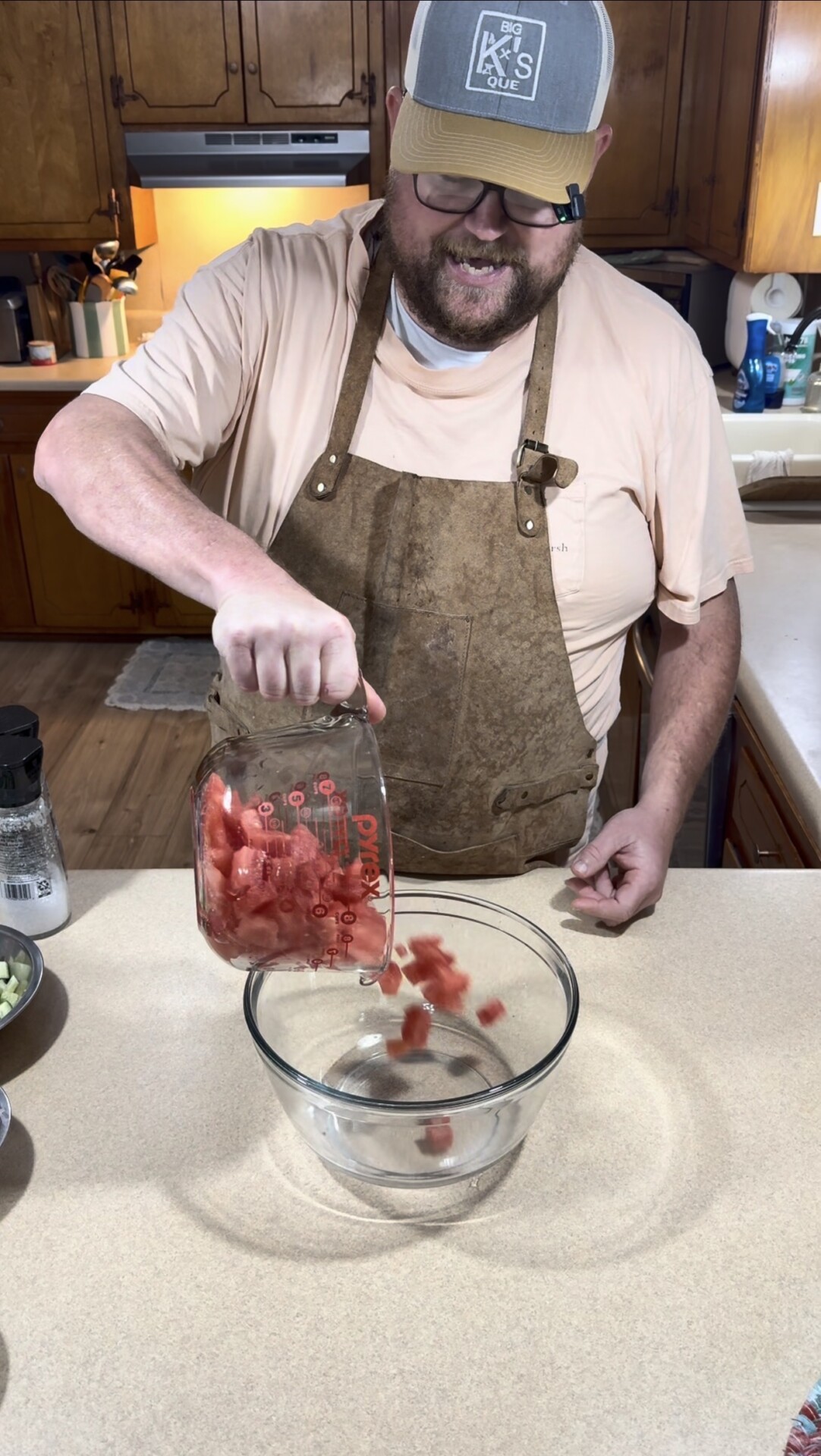 A person wearing an apron and hat pours cubed watermelon from a glass measuring cup into a clear glass bowl on a kitchen counter.