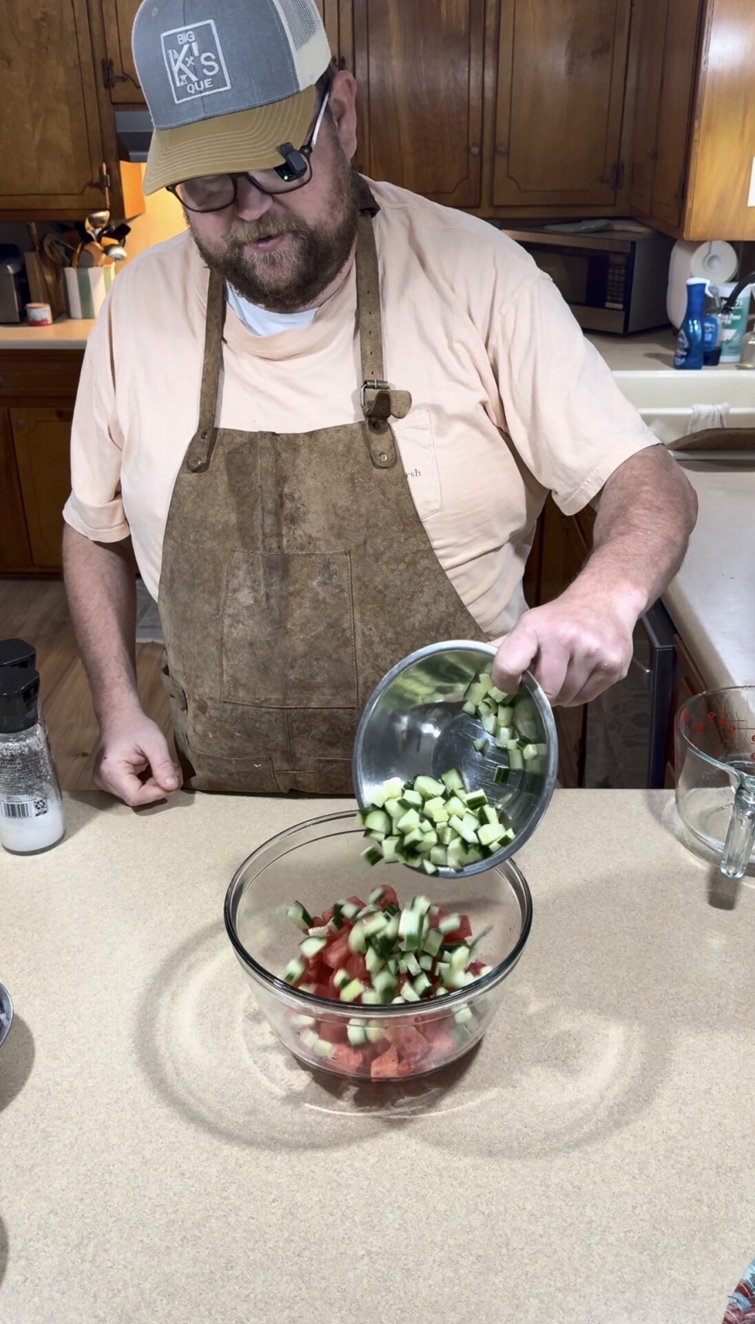 A person wearing a brown apron and cap pours chopped cucumber into a glass bowl of diced watermelon on a kitchen counter.