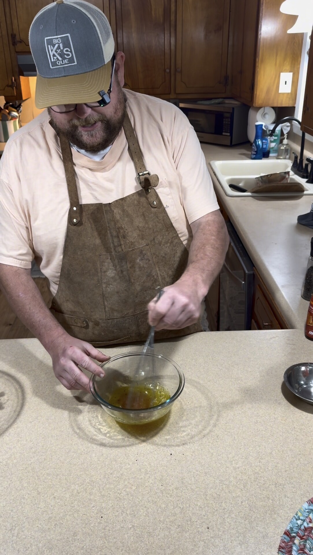 A man wearing an apron and a baseball cap is whisking a yellow mixture in a glass bowl on a kitchen counter. Wooden cabinets, a sink, and kitchen items are visible in the background.